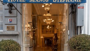 La entrada del Hotel Manos Stephanie con un toldo azul, puertas de cristal y un candelabro en el interior.