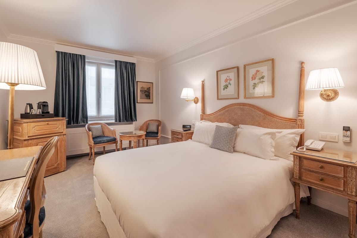 Une chambre d’hôtel avec un grand lit, un bureau avec une lampe, une chaise, une commode et une fenêtre avec des rideaux.