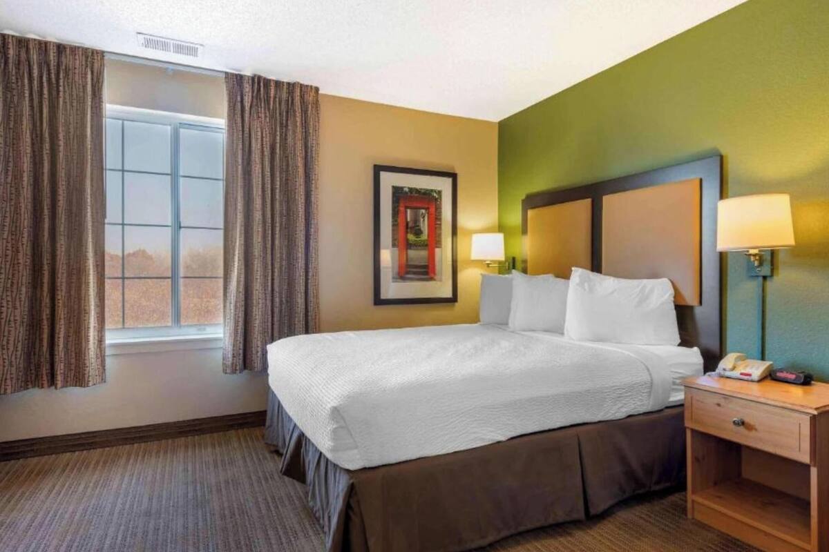 Une chambre d’hôtel avec un lit, une table de chevet, une lampe, un tableau encadré au mur et une fenêtre avec des rideaux.