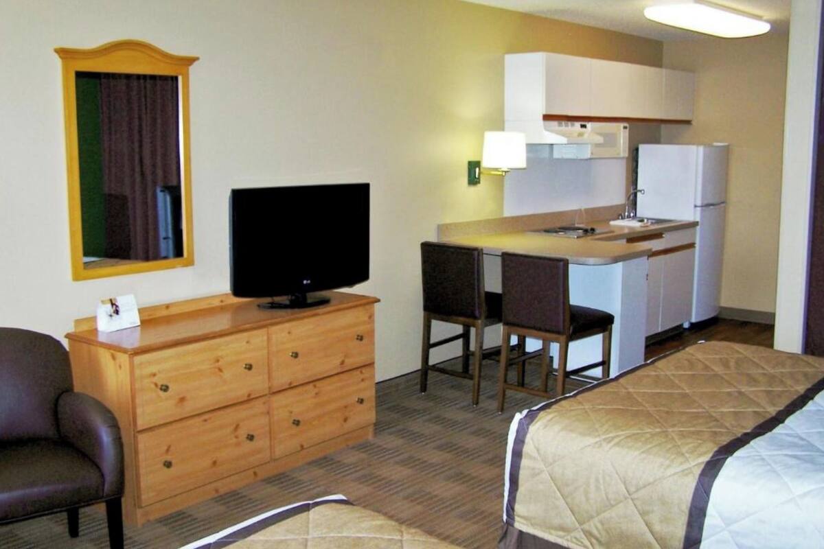 Une chambre d’hôtel avec une kitchenette, un téléviseur posé sur une commode en bois, un lit avec une courtepointe et une chaise.
