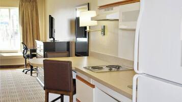 A compact hotel room with a kitchenette, a dining area, a flat-screen TV, a window with curtains, and a carpeted floor.