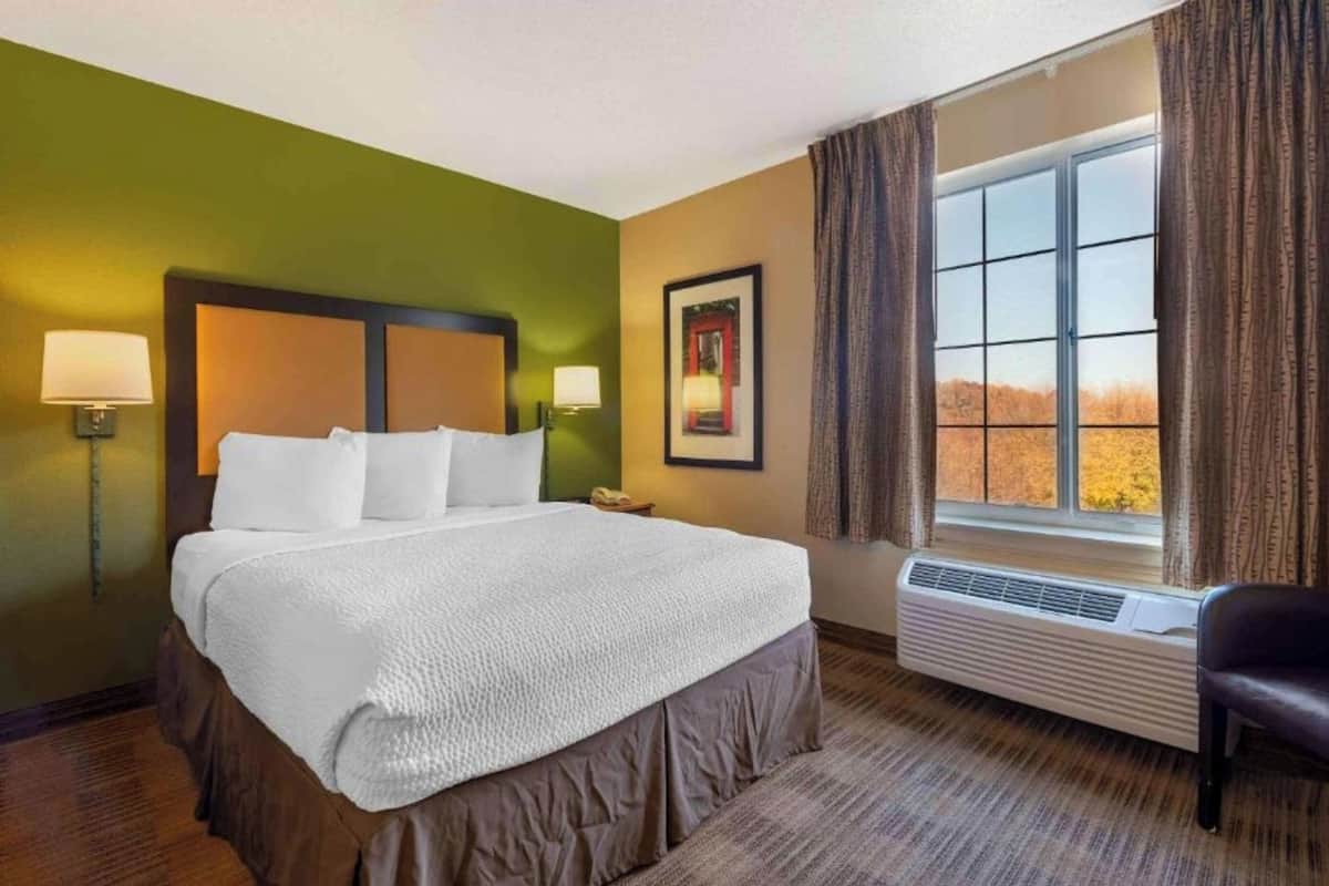 Une chambre d’hôtel avec un lit, deux lampes de chevet, une fenêtre avec des rideaux, un tableau encadré au mur et une chaise.