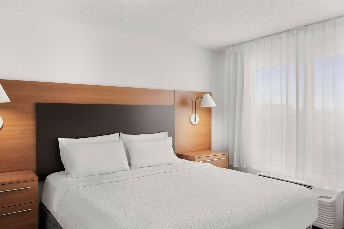 A hotel room with a large bed, a wooden headboard, a bedside table with a lamp, and a window with sheer curtains.