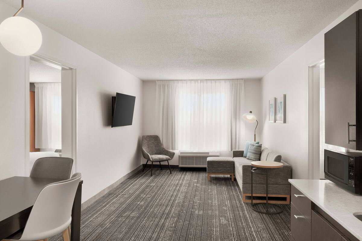 A modern hotel room with a flat-screen TV, a dining area with a table and chairs, a sofa, a small round table, and a kitchenette.