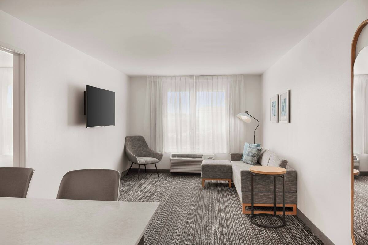 A hotel room with a flat-screen TV mounted on the wall, a sofa with a side table, a chair, and a dining table.