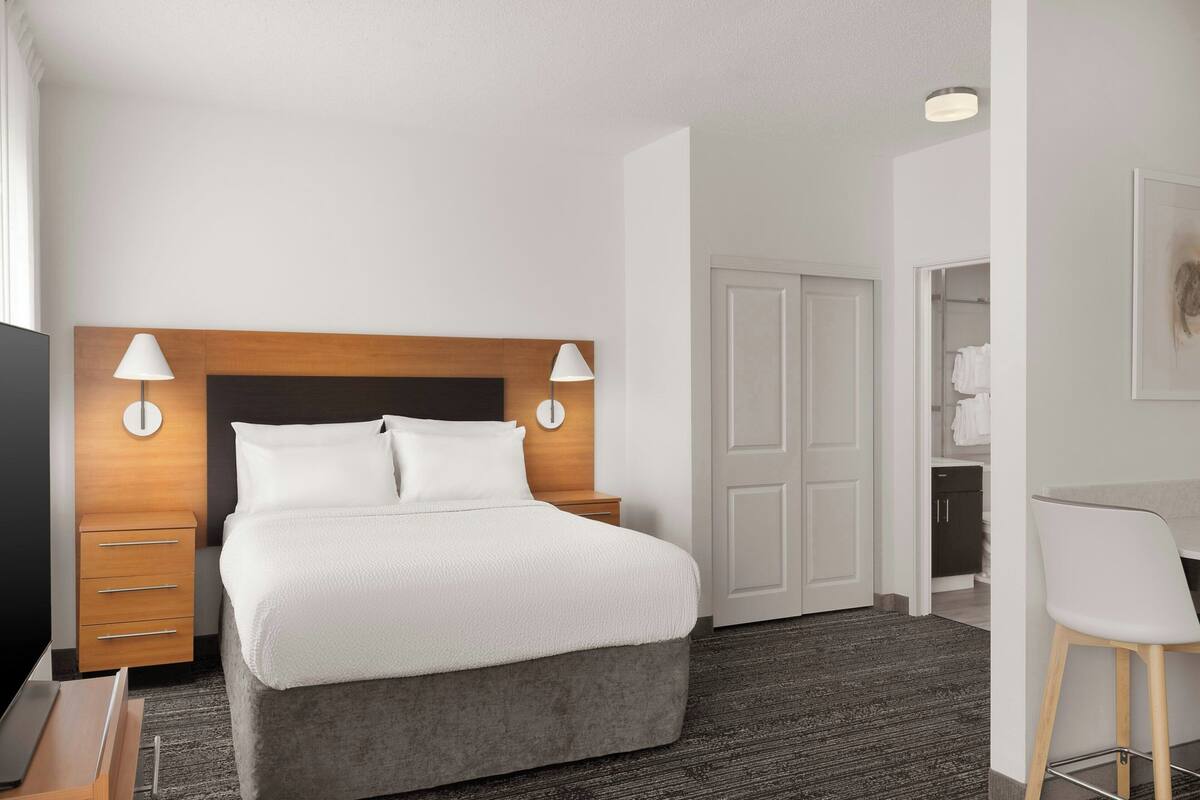 A hotel room with a bed, bedside lamps, a wooden headboard, a desk with a lamp, a chair, and a bathroom visible through an open door.