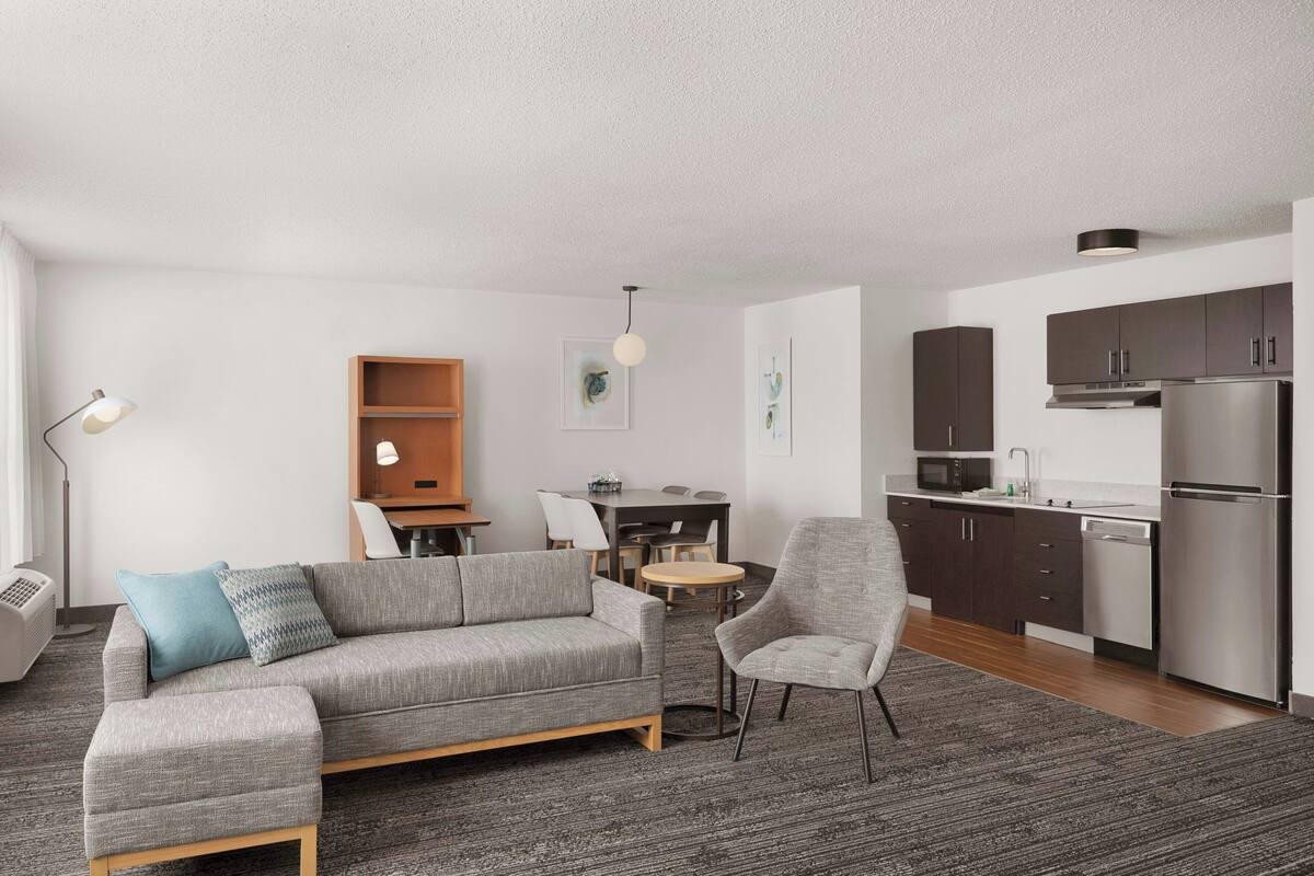 A modern living room with a grey sectional sofa, a grey armchair, and a small round table. There is a kitchen area with dark cabinets and stainless steel appliances.