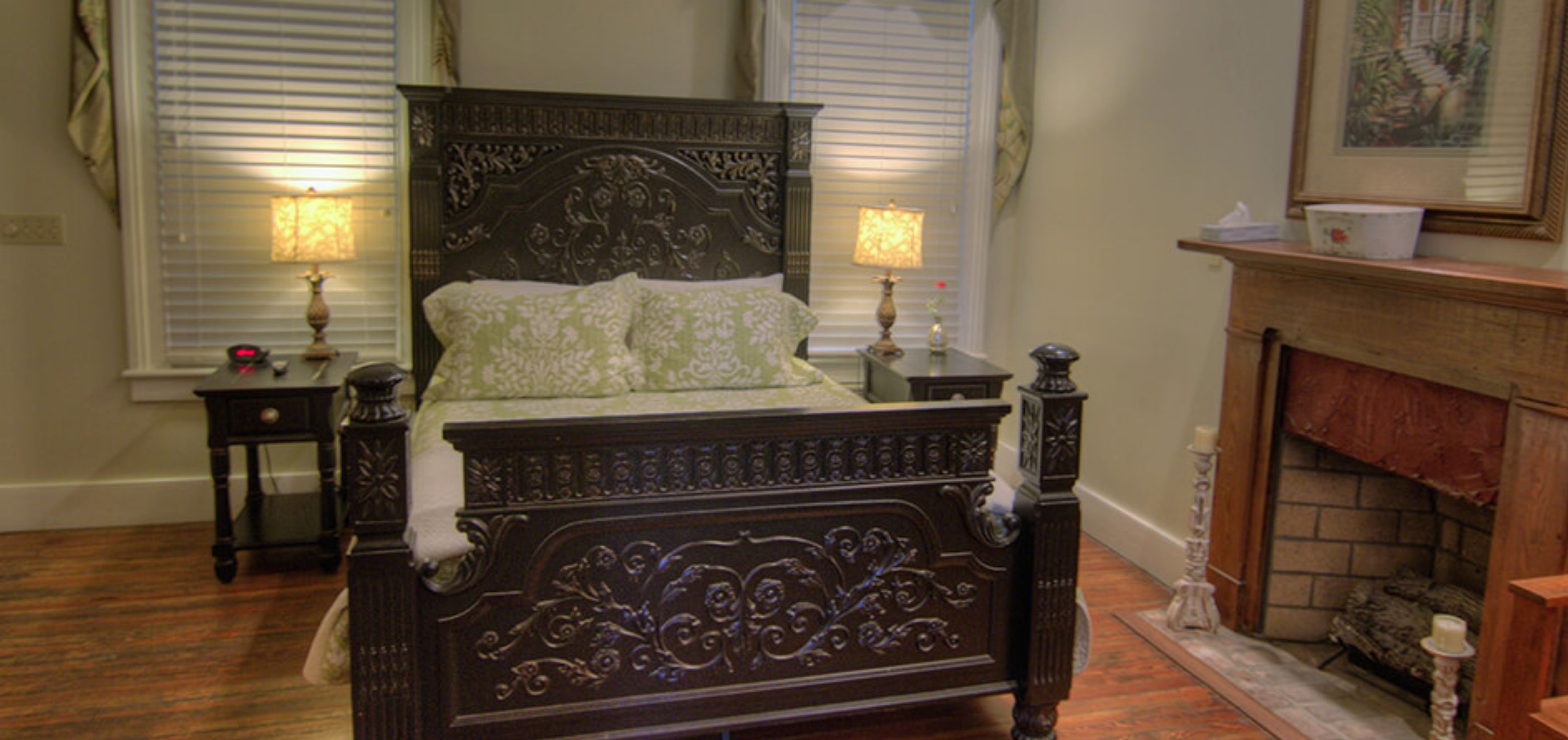 A bedroom with a large wooden bed, two bedside tables with lamps, a fireplace, and a window with blinds.