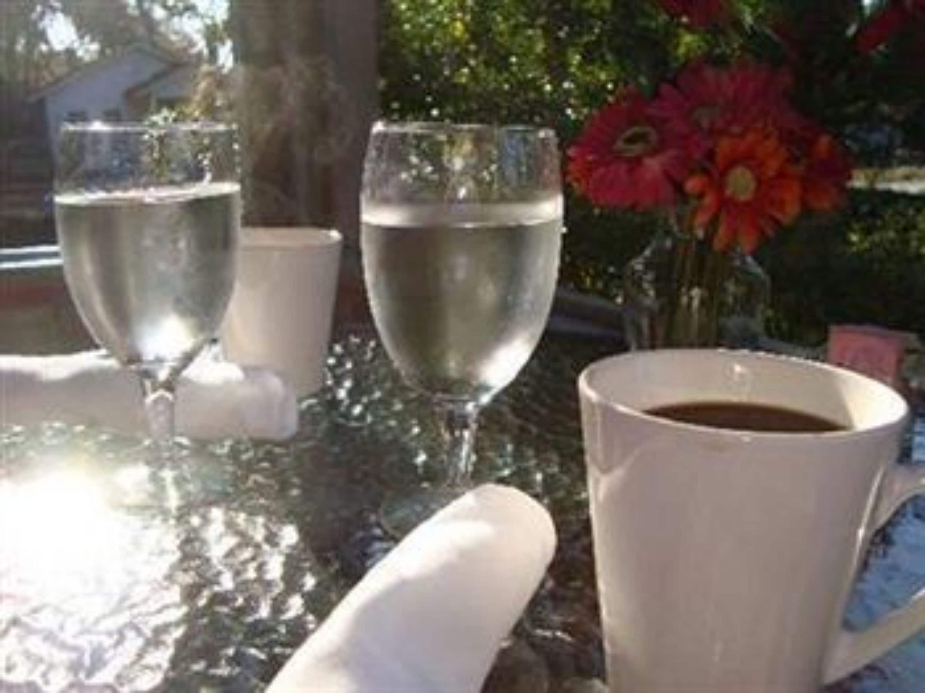 Una mesa con dos vasos de agua y una taza blanca, posiblemente de café, con un jarrón de flores rojas al fondo.