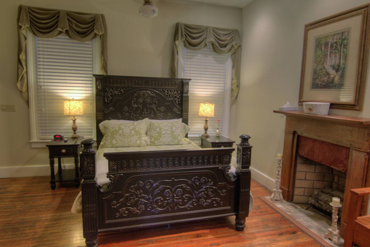 A traditional bedroom with a four-poster bed, two bedside tables, a fireplace, and two windows with curtains.