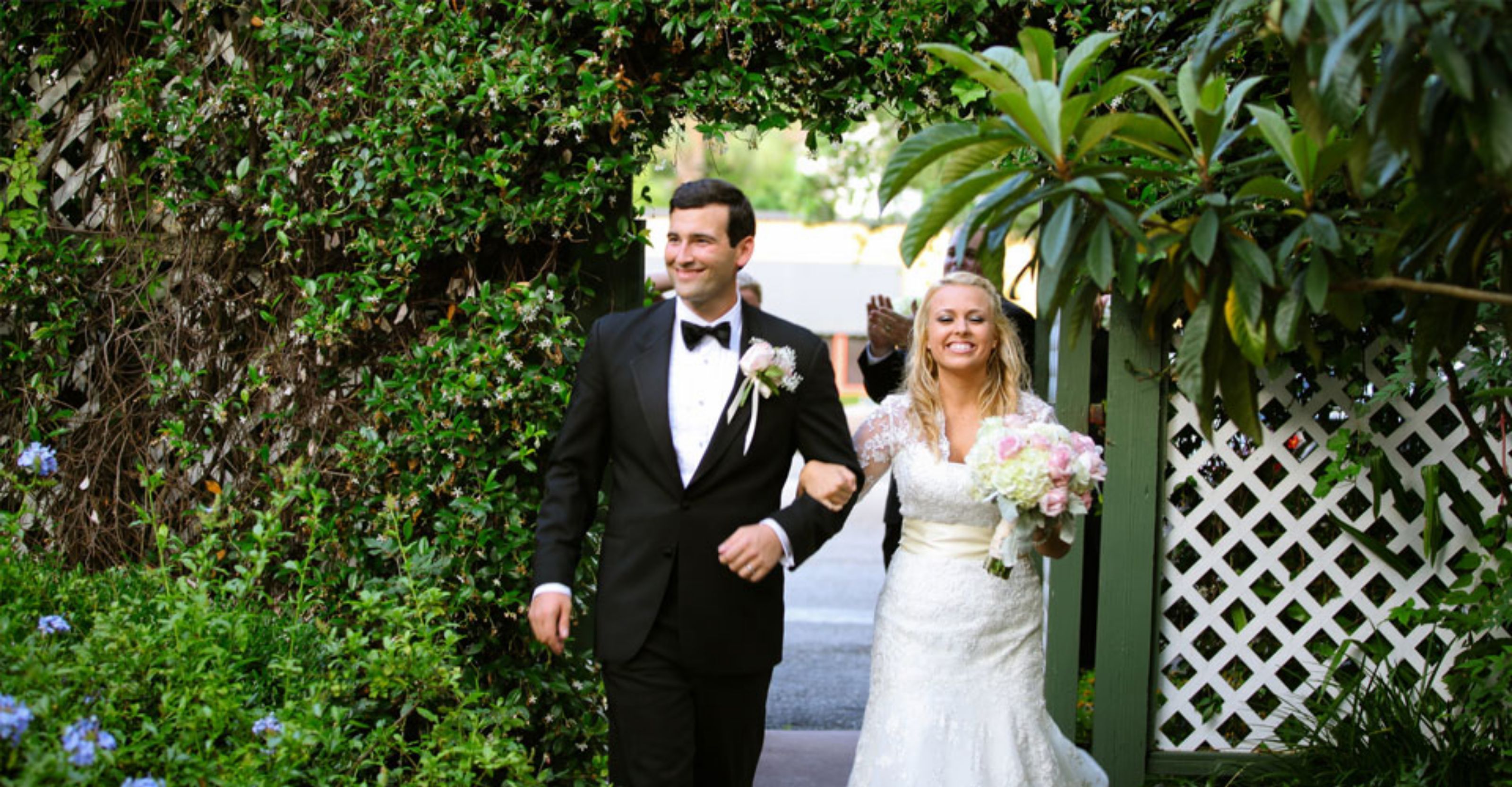 A couple walking through a garden with a white lattice fence and lush greenery.
