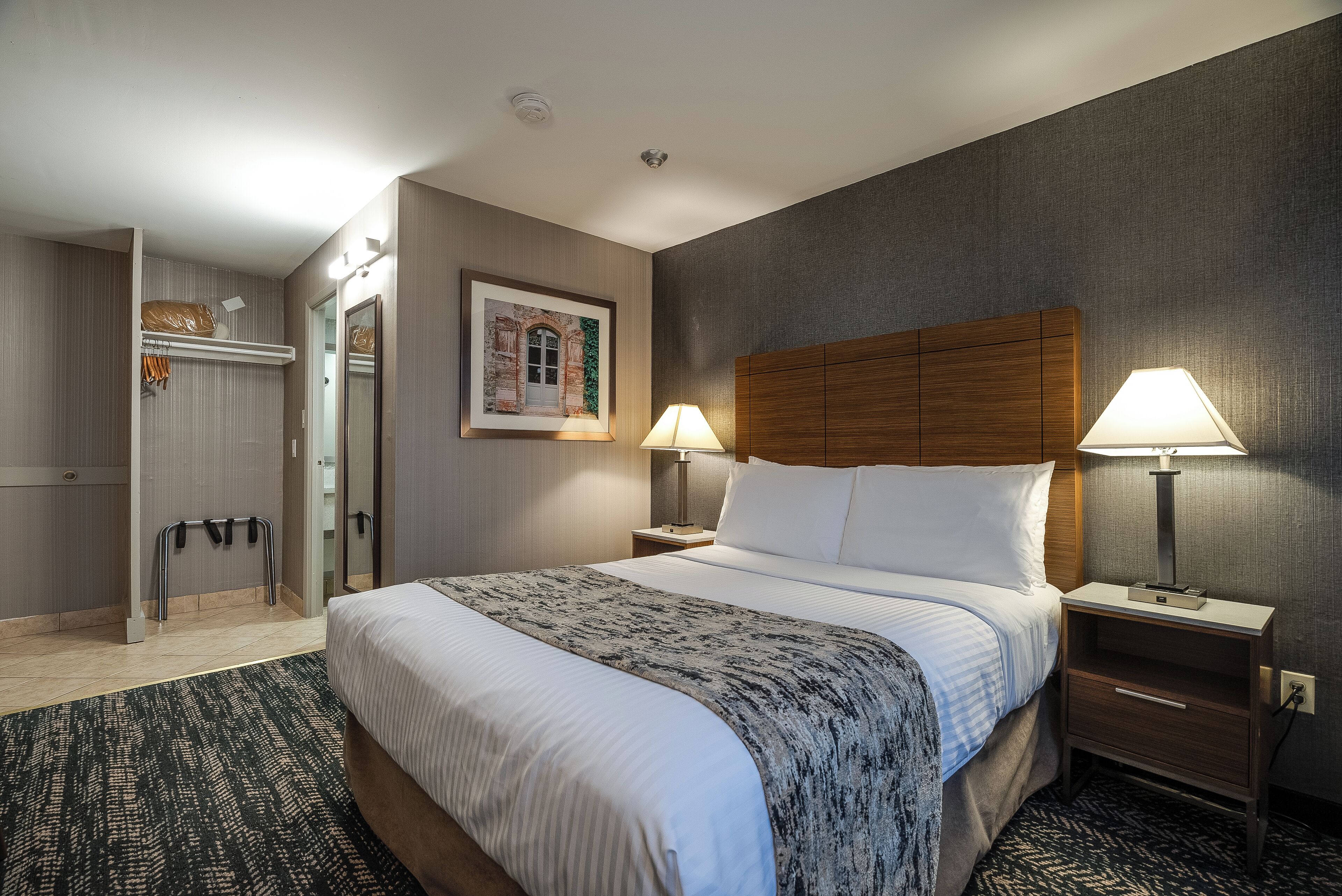 Habitación de hotel con una cama grande, dos mesitas de noche con lámparas, un cabecero de madera, un cuadro enmarcado en la pared y un perchero.