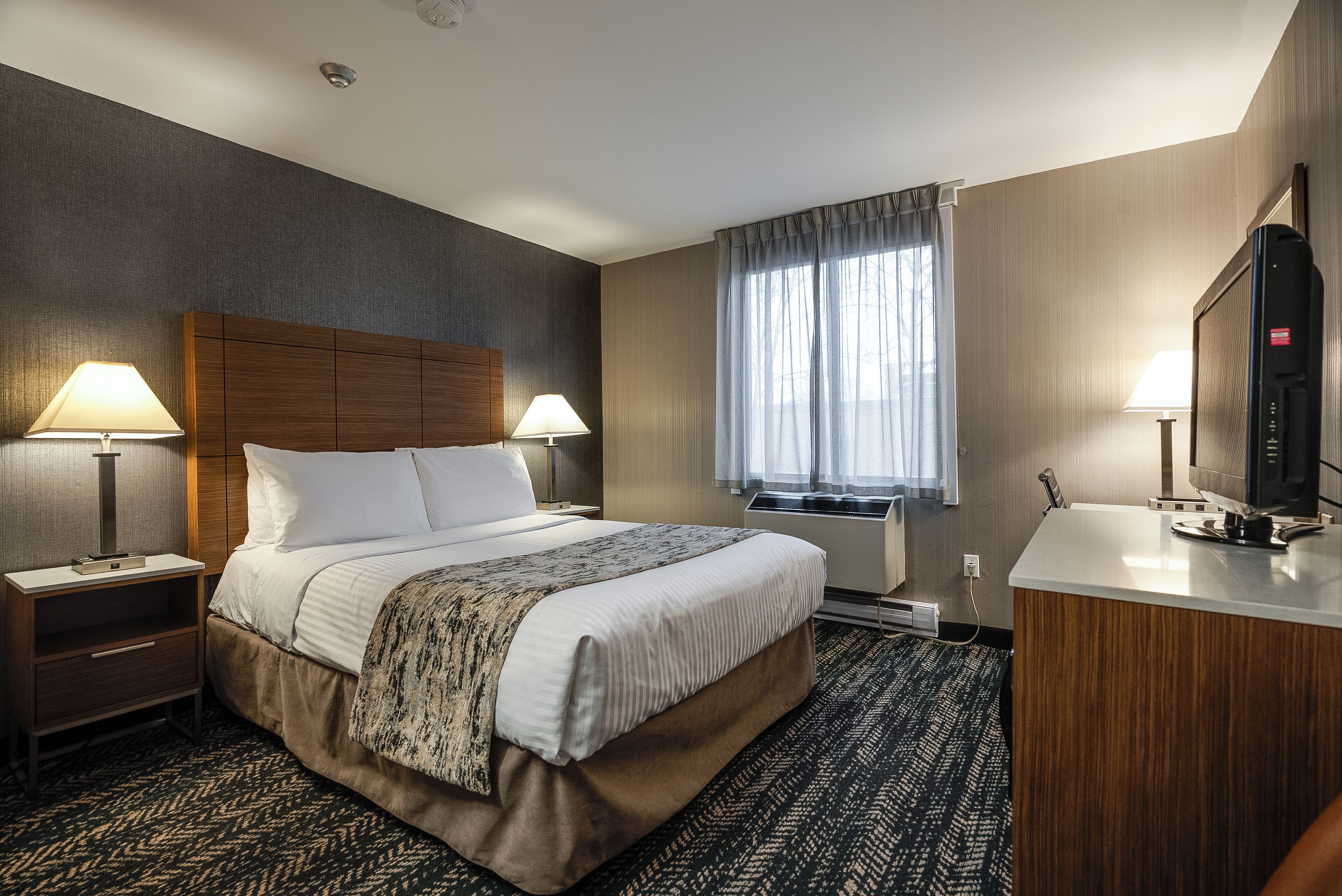 A hotel room with a large bed, two bedside tables with lamps, a TV, and a window with curtains.