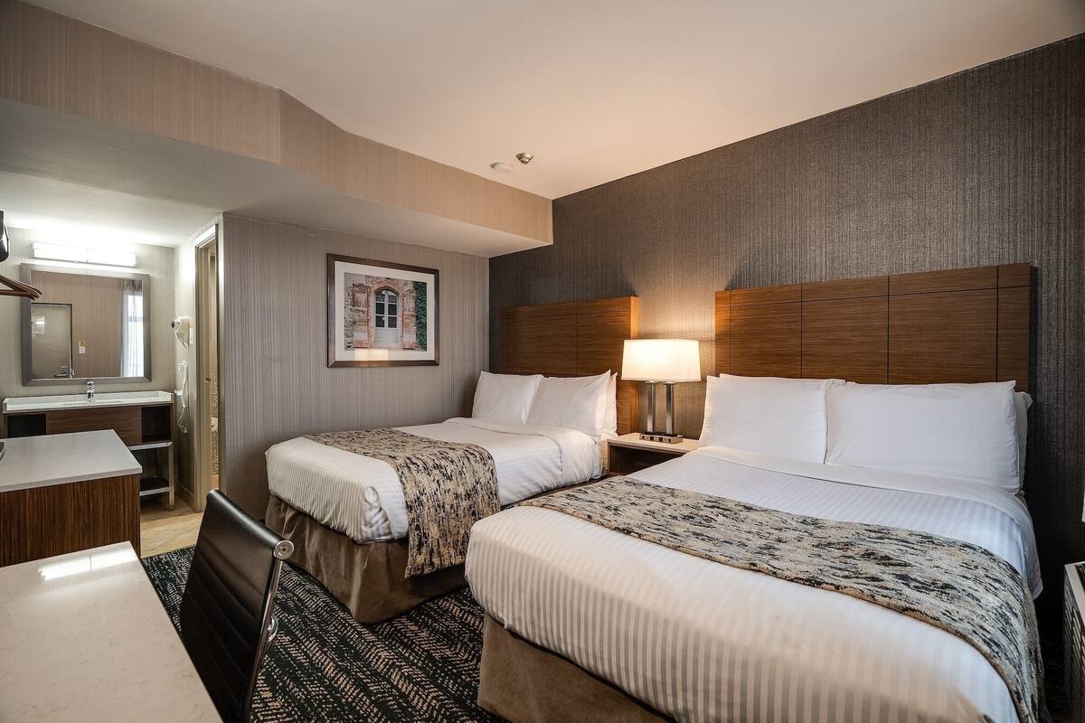 A hotel room with two beds, a bedside table with a lamp, a bathroom with a mirror and sink, and a framed picture on the wall.