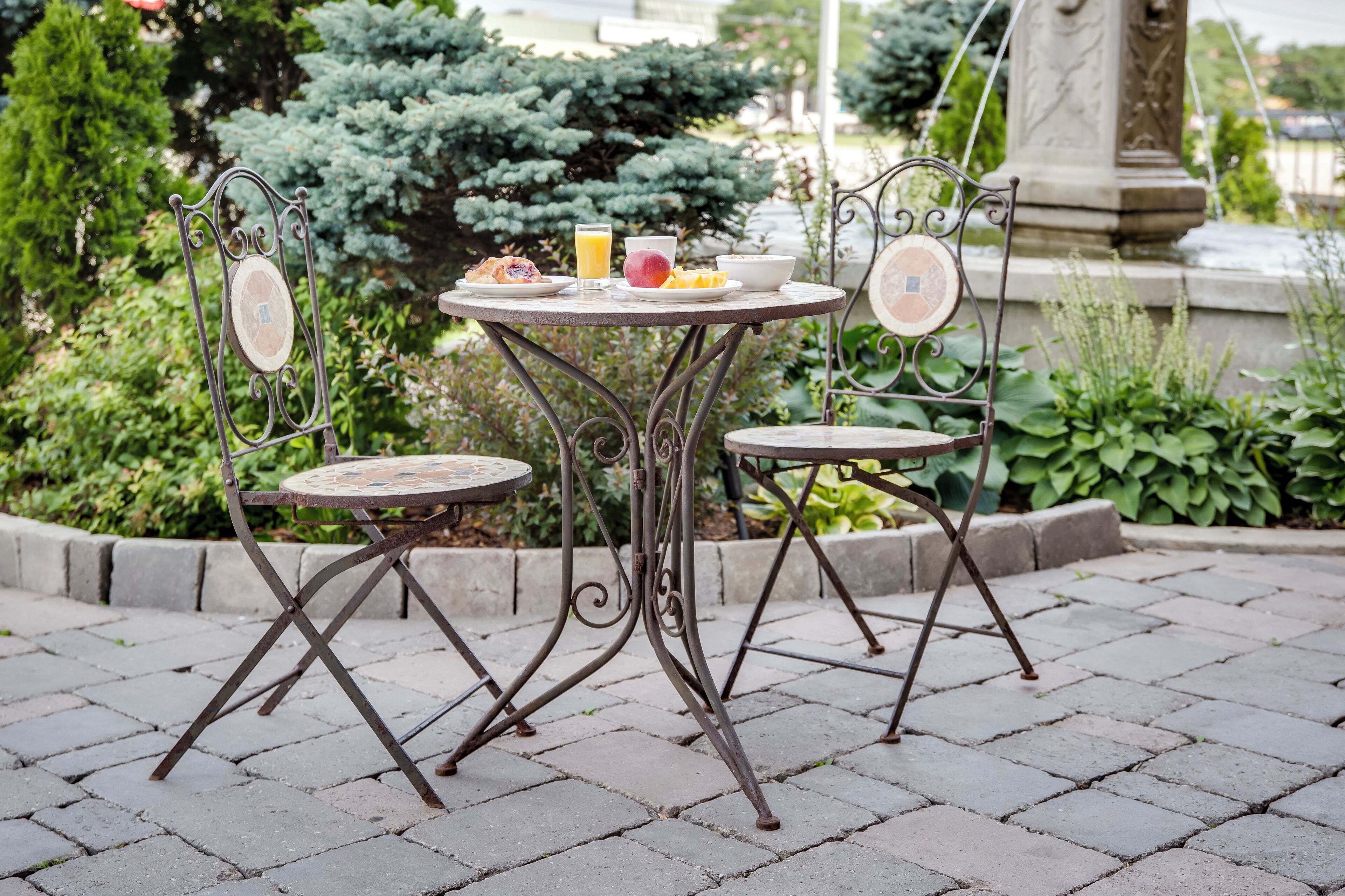 Une table ronde en métal, entourée de chaises, se trouve au milieu d’un jardin où poussent différentes plantes, et une fontaine en pierre est visible au fond.