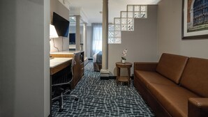 A hotel room with a bed, a desk with a lamp, a brown sofa, a patterned carpet, and a framed picture on the wall. - Monte Carlo Inn Toronto West Suites (Mississauga)