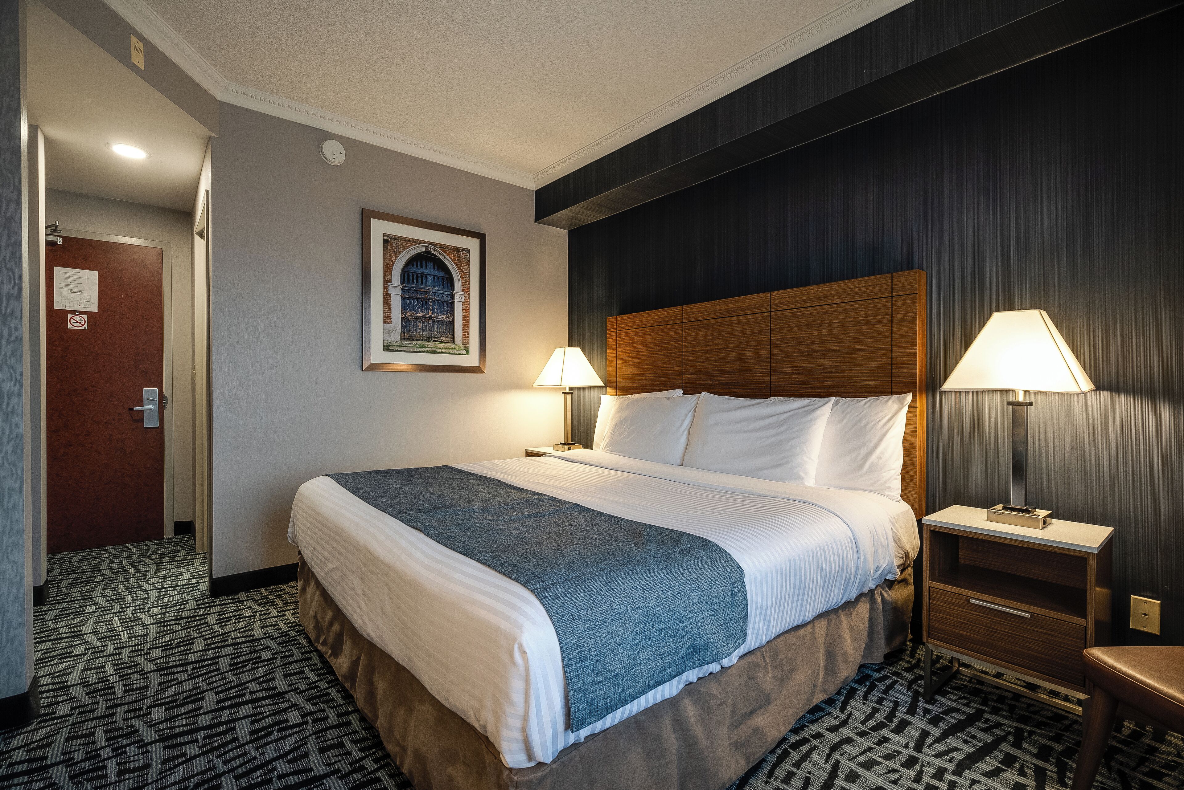 A hotel room with a large bed, two bedside tables with lamps, a framed picture on the wall, and a patterned carpet.