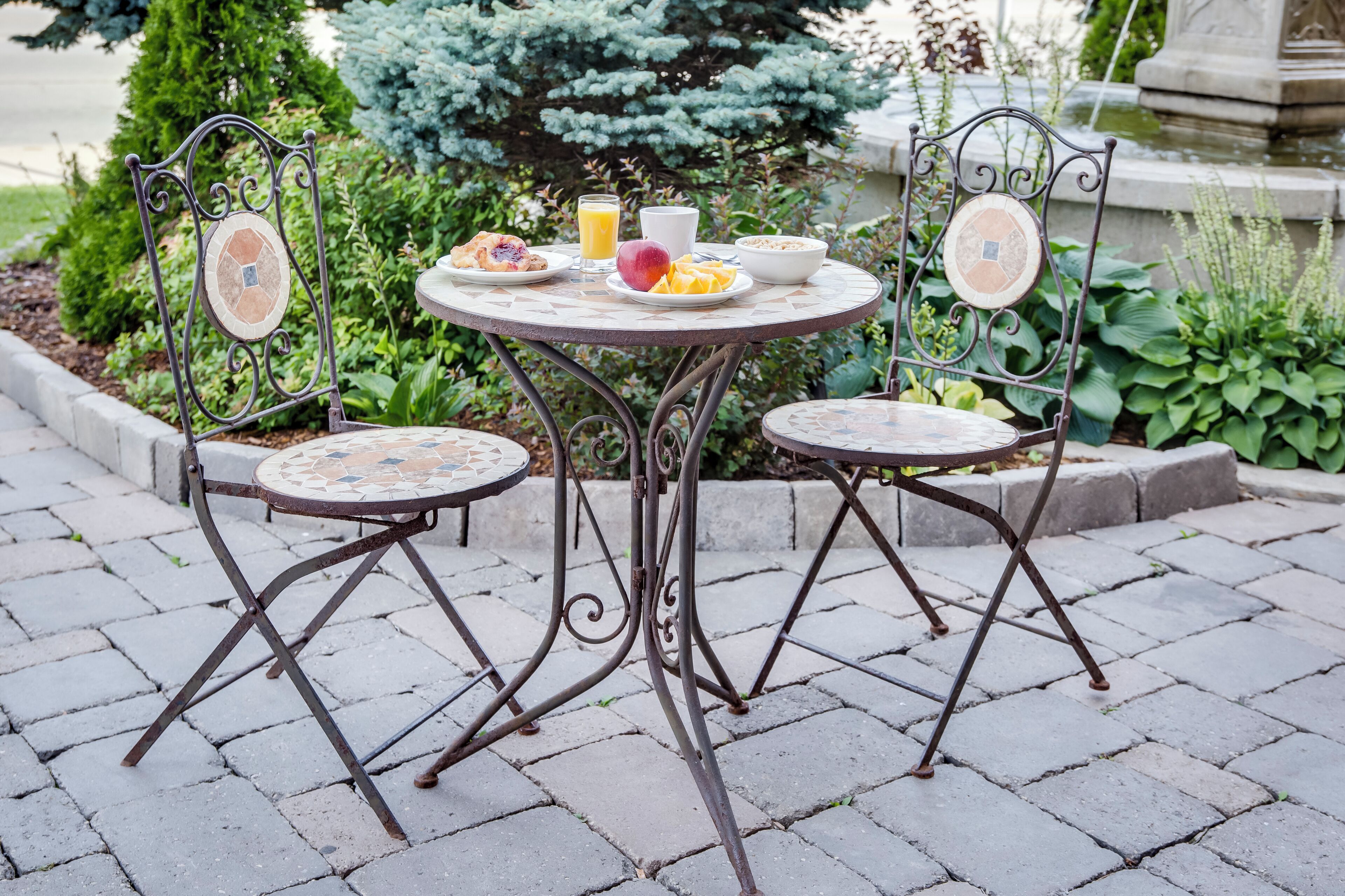 Een ronde metalen tafel met twee stoelen, omgeven door een tuin met planten en een fontein op de achtergrond.