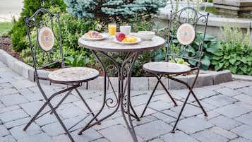 A round metal table with two chairs, surrounded by a garden with plants and a fountain in the background.