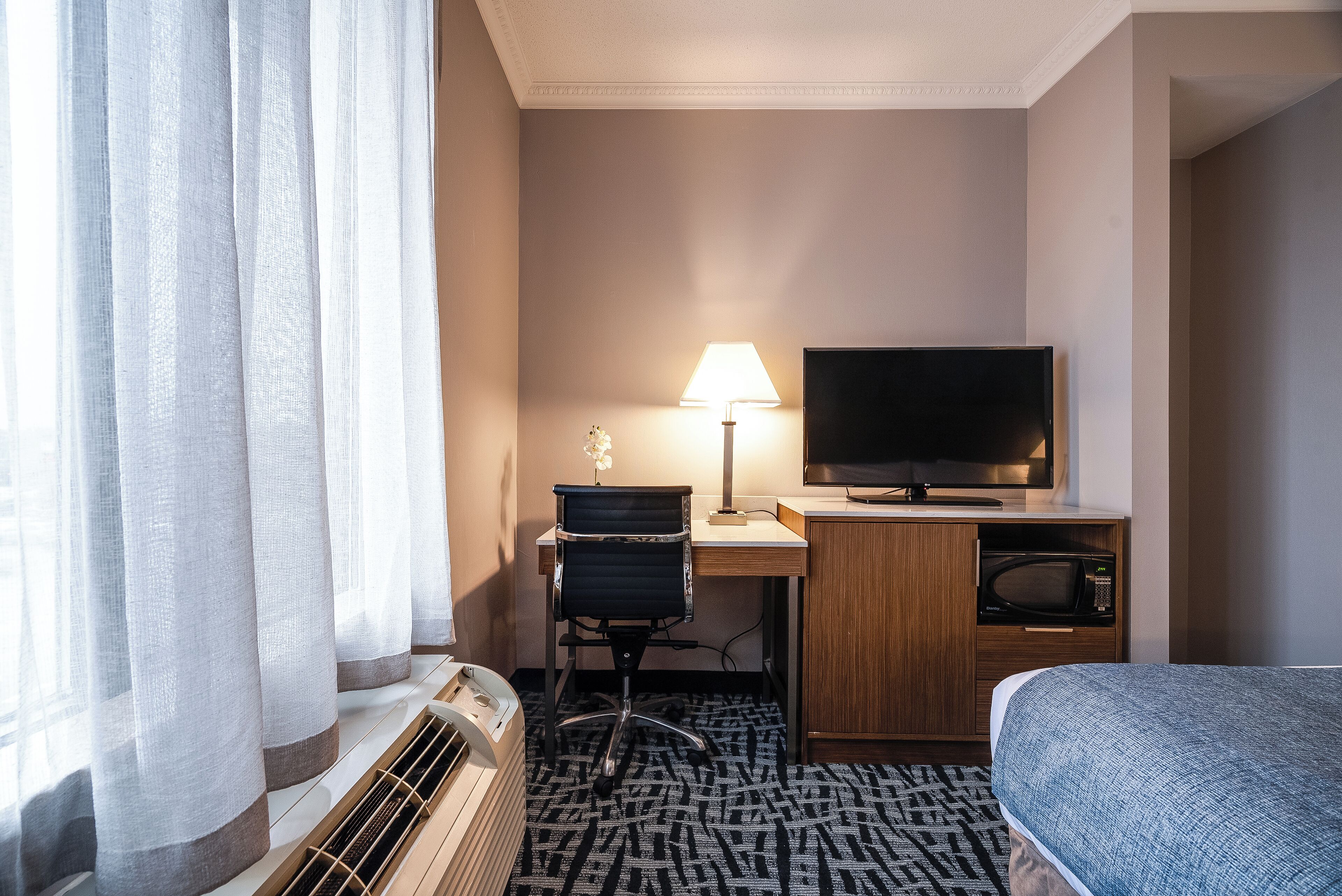 A hotel room with a bed, a desk with a chair, a television, a microwave, and a window with curtains.