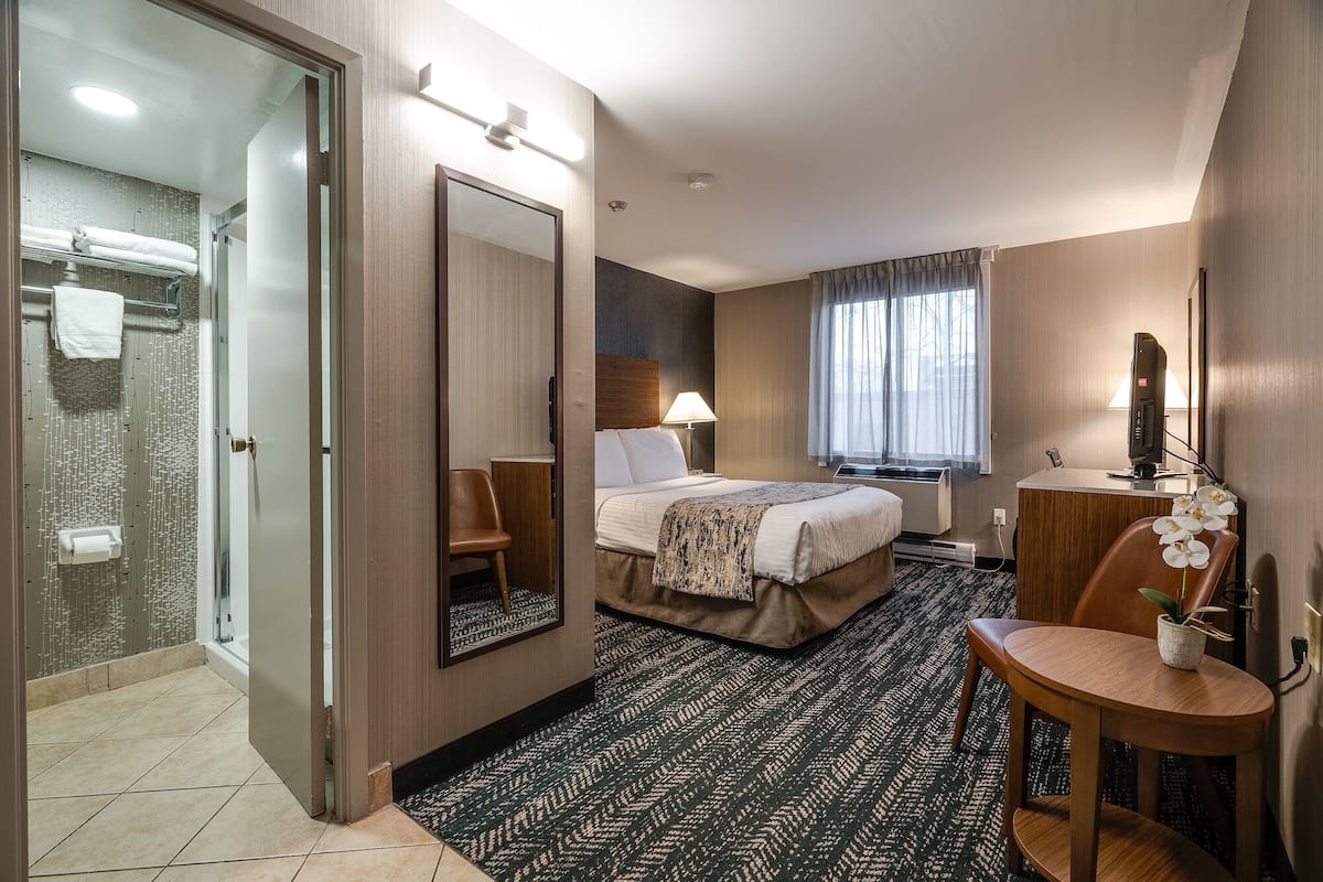 A hotel room with a bed, a desk, a chair, a mirror, a shower, and a window with curtains.