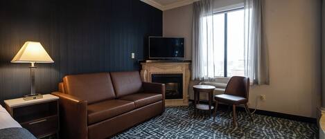 Une chambre d’hôtel avec un canapé en cuir marron, un téléviseur à écran plat, une cheminée, une petite table ronde avec une chaise et une moquette à motifs.