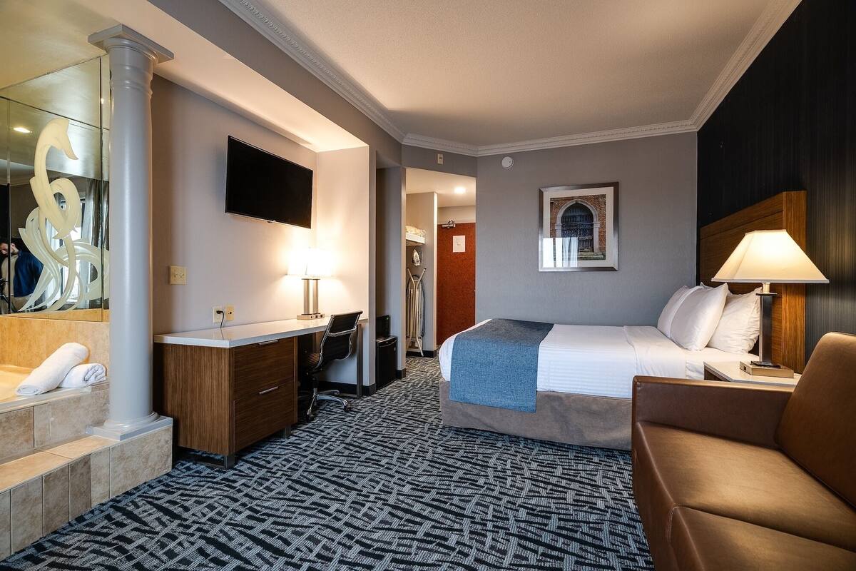 A hotel room with a bed, a desk with a chair, a bathroom with a towel, and a mirror.