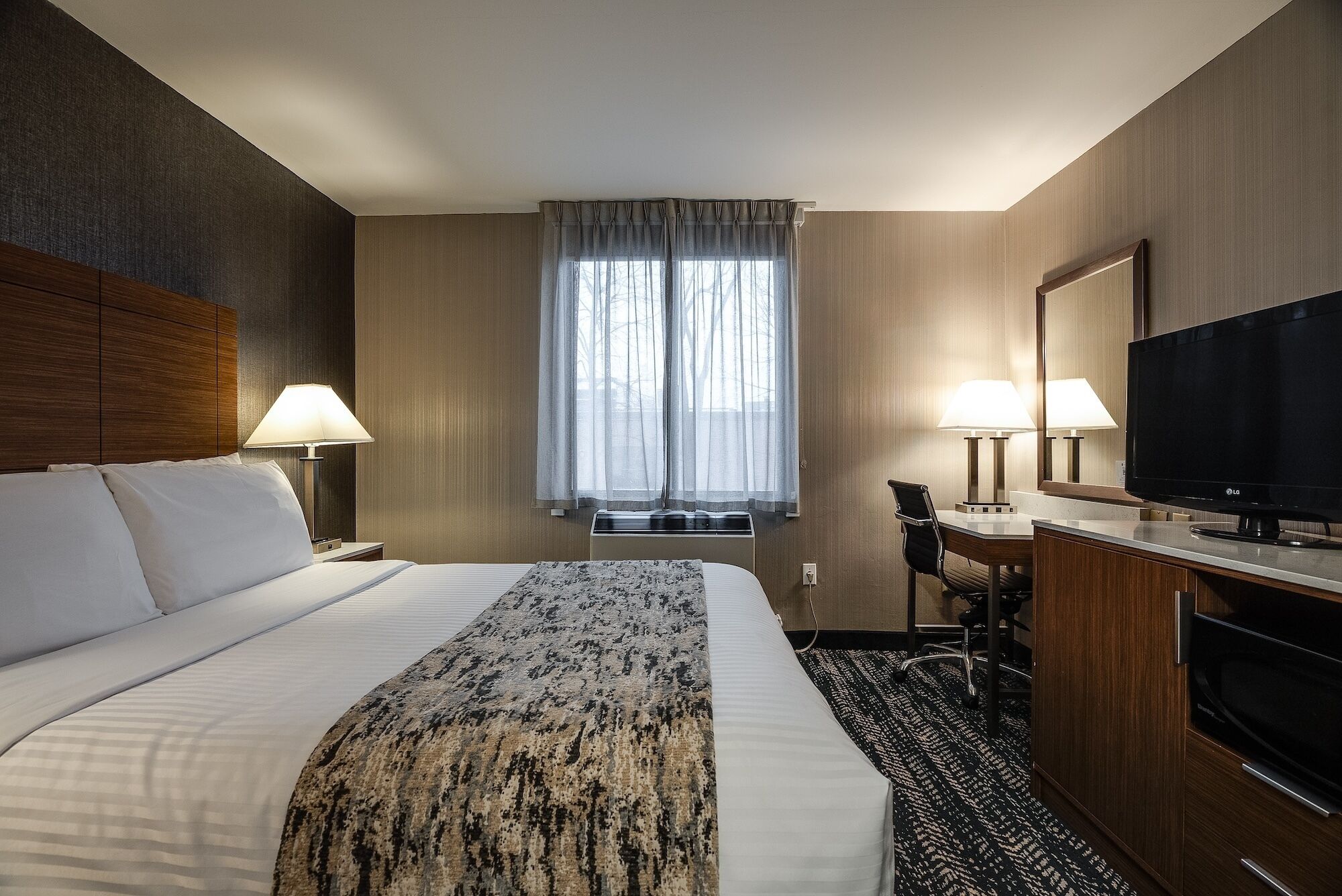 A hotel room with a large bed, a desk with a chair, a television, and a window with curtains.