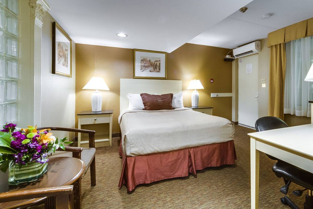 A hotel room with a bed, two bedside tables with lamps, a desk with a chair, a small table with a vase of flowers, and an air conditioning unit.