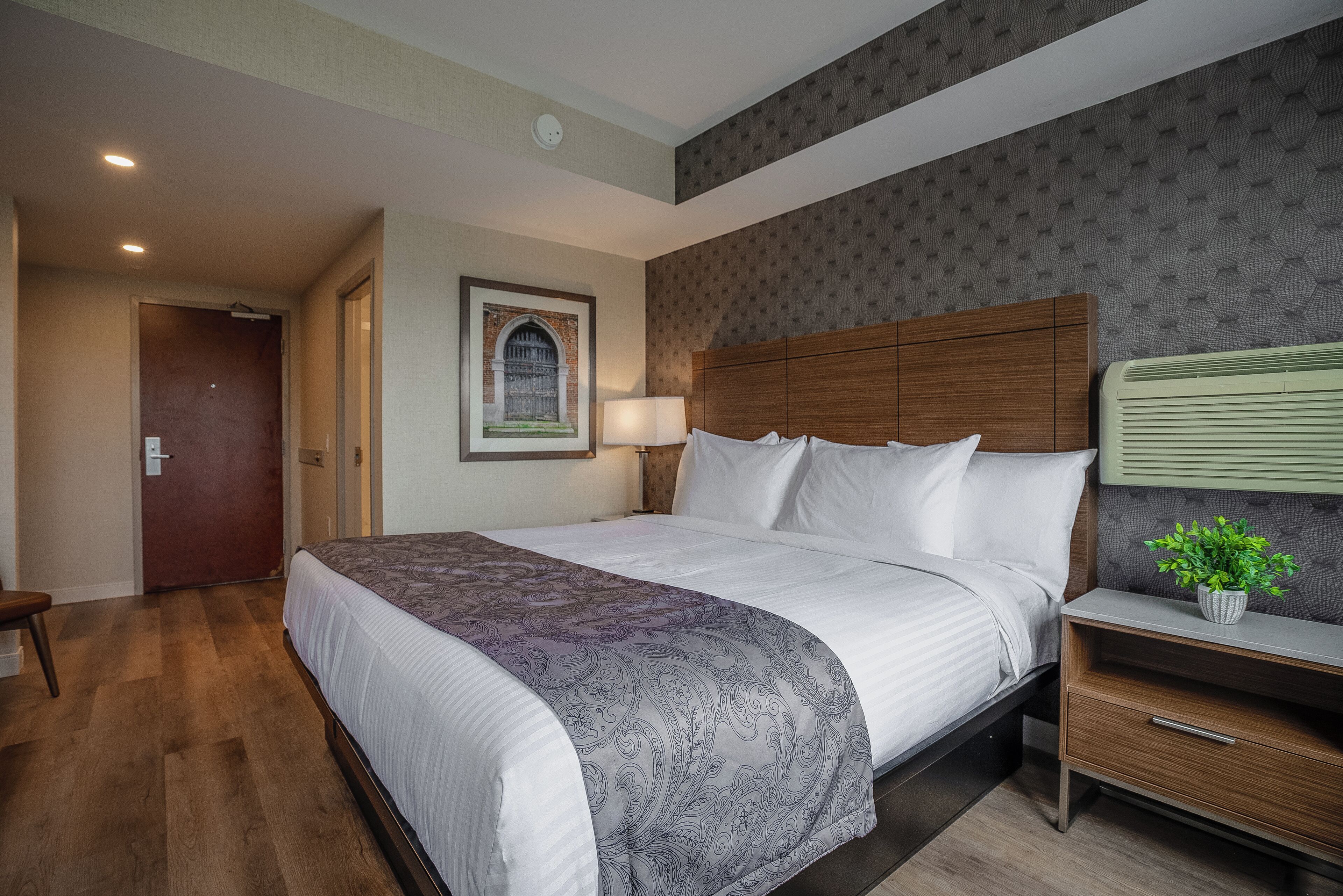 A hotel room with a large bed, a wooden nightstand, a bedside lamp, and a framed picture on the wall.