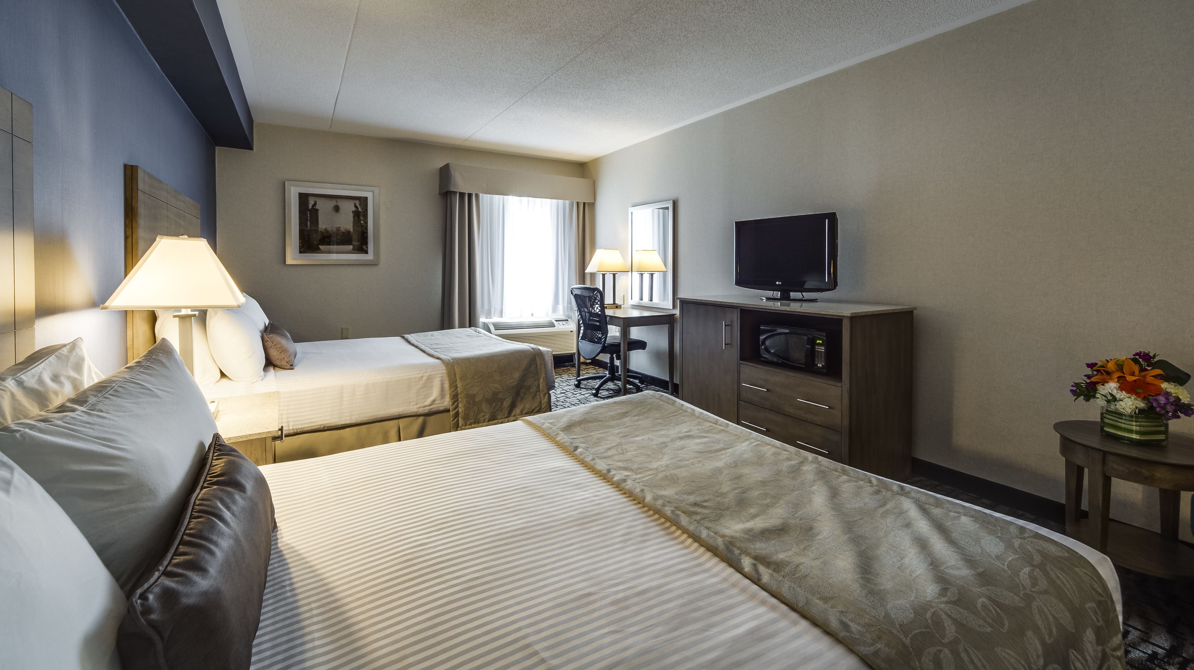 Habitación de hotel con dos camas, un escritorio con televisor, microondas y una pequeña mesa con un florero.