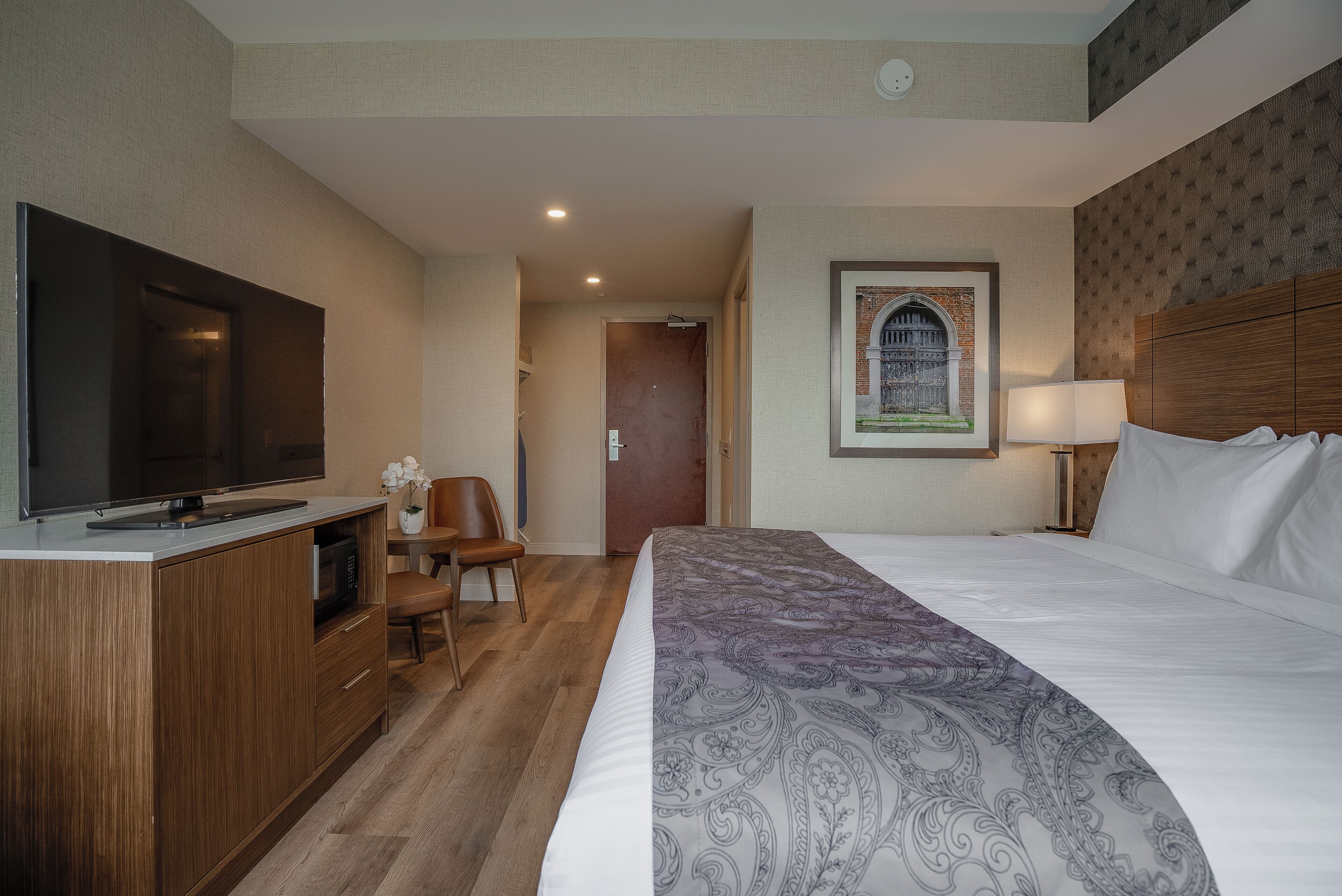 Ein Hotelzimmer mit einem großen Bett, einem Fernseher auf einer Holzkommode, einem Schreibtisch mit Stuhl und einem gerahmten Bild an der Wand.