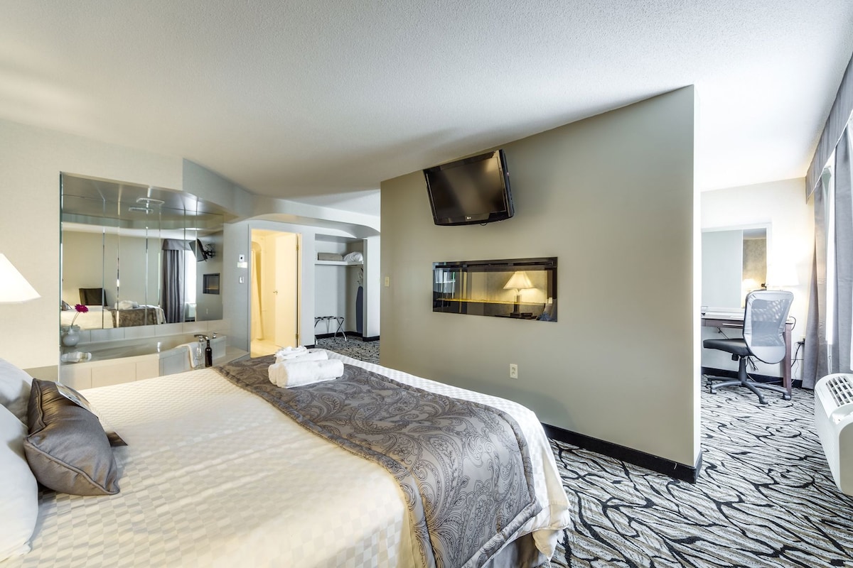 A hotel room with a bed, a desk, a chair, a TV, a mirror, and a bathroom.