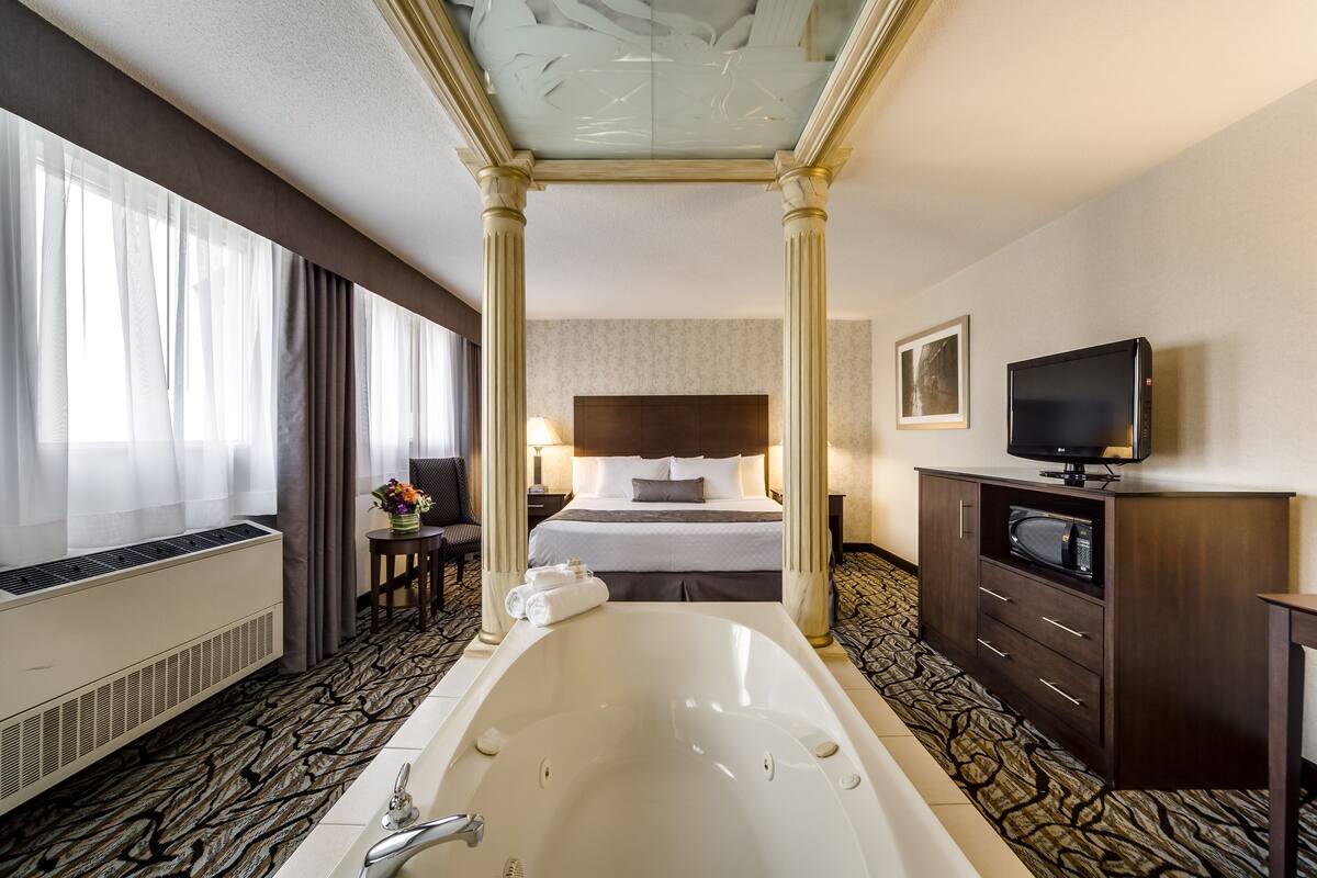 Une chambre d’hôtel dotée d’une grande baignoire, d’un lit avec une tête de lit sombre, d’un téléviseur posé sur un meuble en bois et d’une fenêtre ornée de rideaux légers.