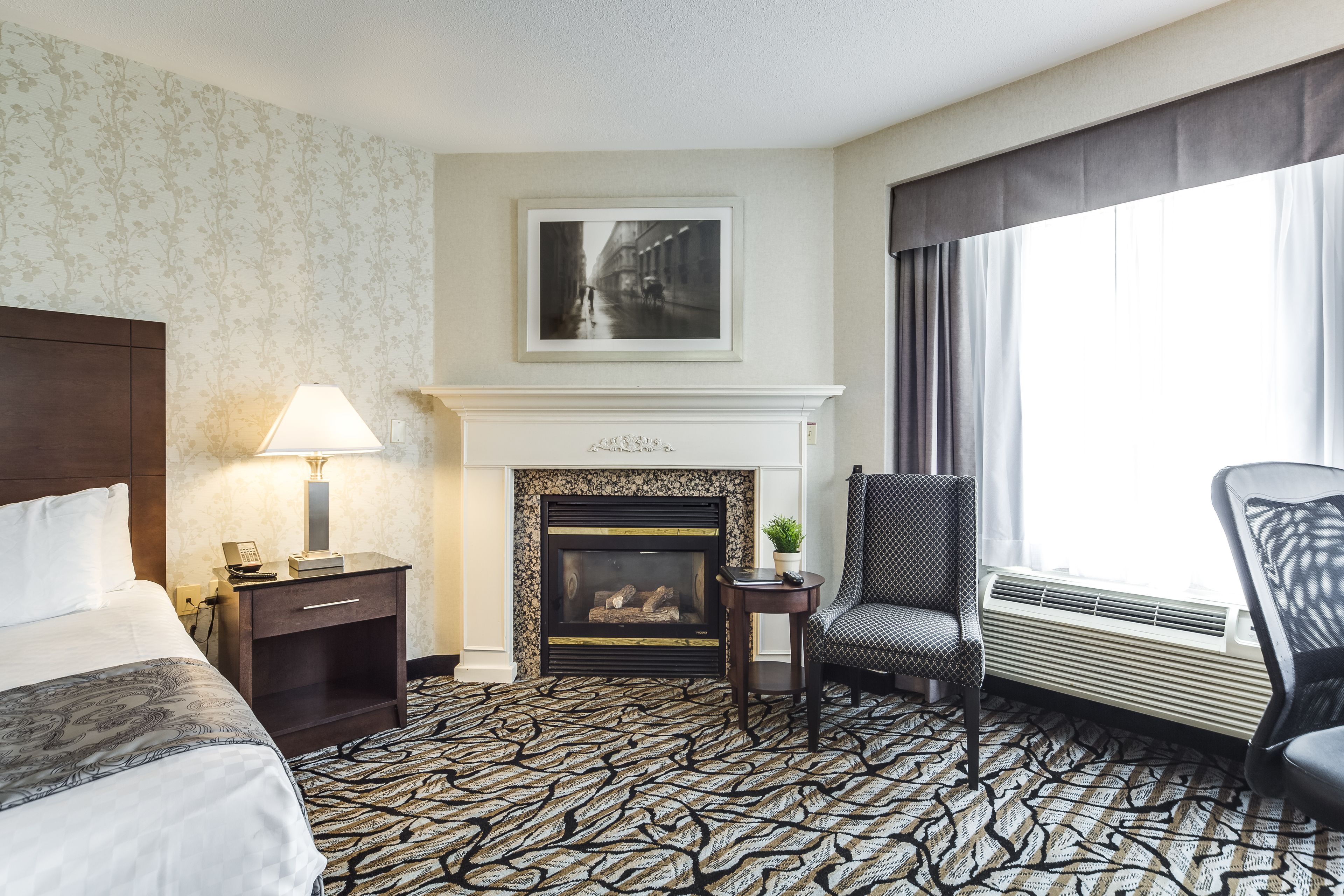 Une chambre d’hôtel avec un lit, une table de chevet avec une lampe, une cheminée, un fauteuil et une grande fenêtre.