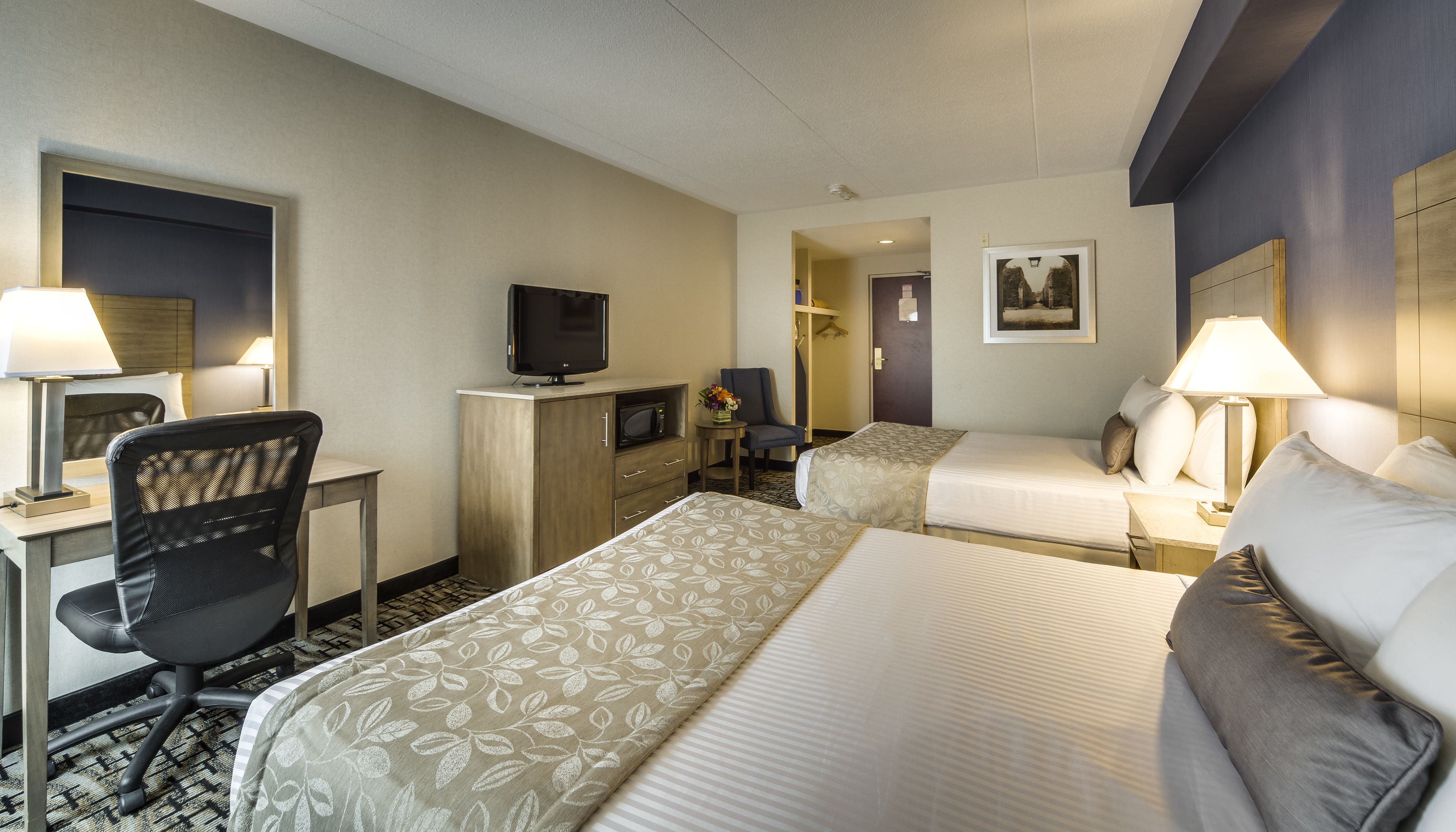 Habitación de hotel con dos camas, un escritorio con una silla, un televisor y una pequeña mesa con un jarrón de flores.