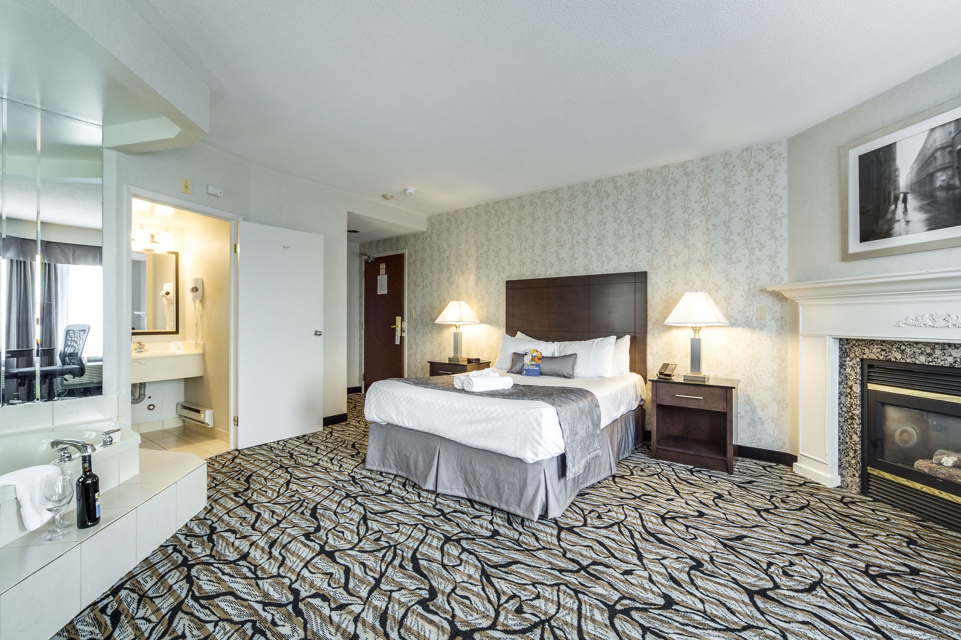 Ein Hotelzimmer mit einem großen Bett, zwei Nachttischlampen, einem Kamin, einem Spiegel und einem Badezimmer mit Waschbecken und Spiegel.