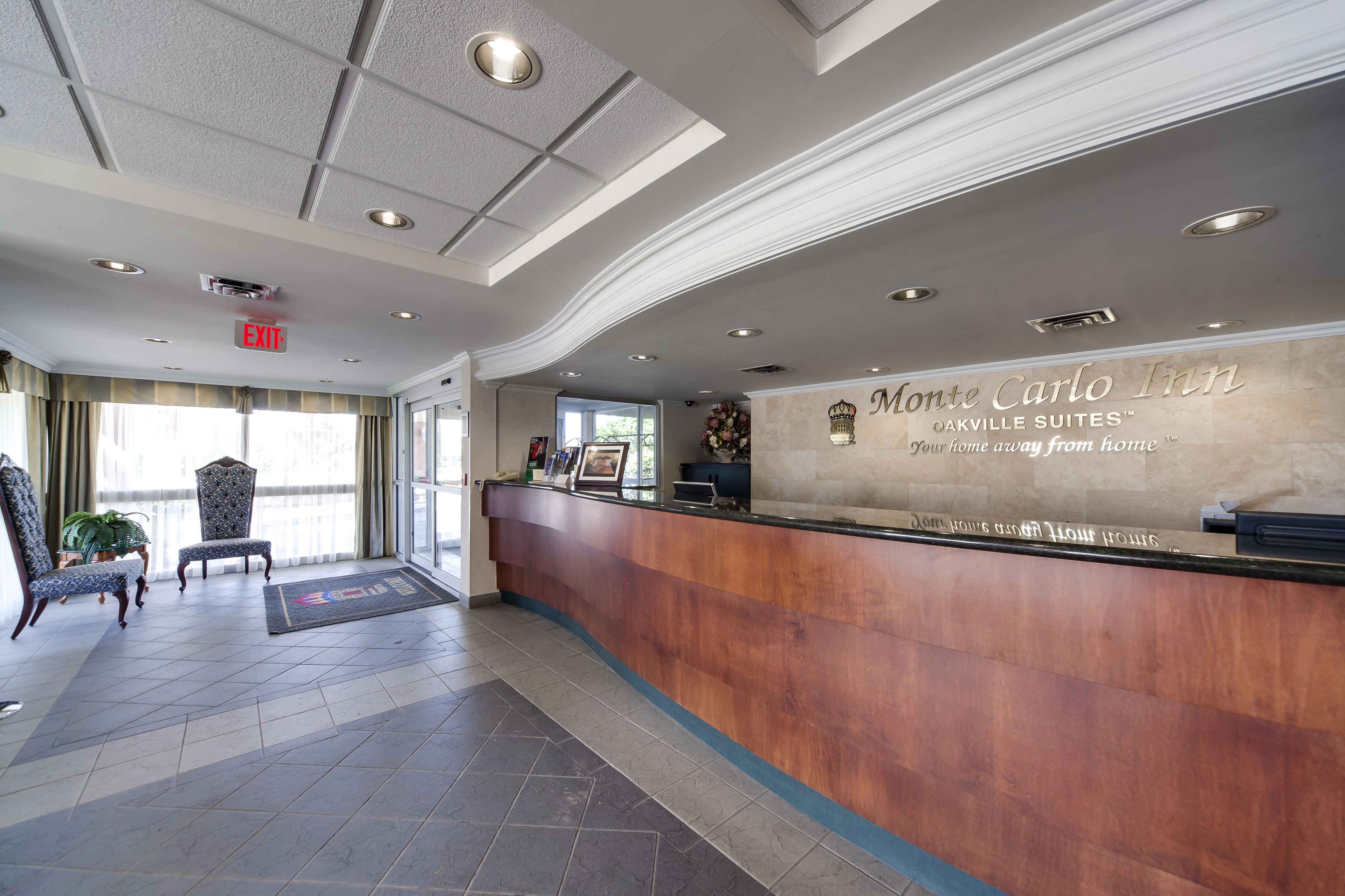 A reception area with a wooden reception desk, a sign for Monte Carlo Inn Oakville Suites, and two upholstered chairs.