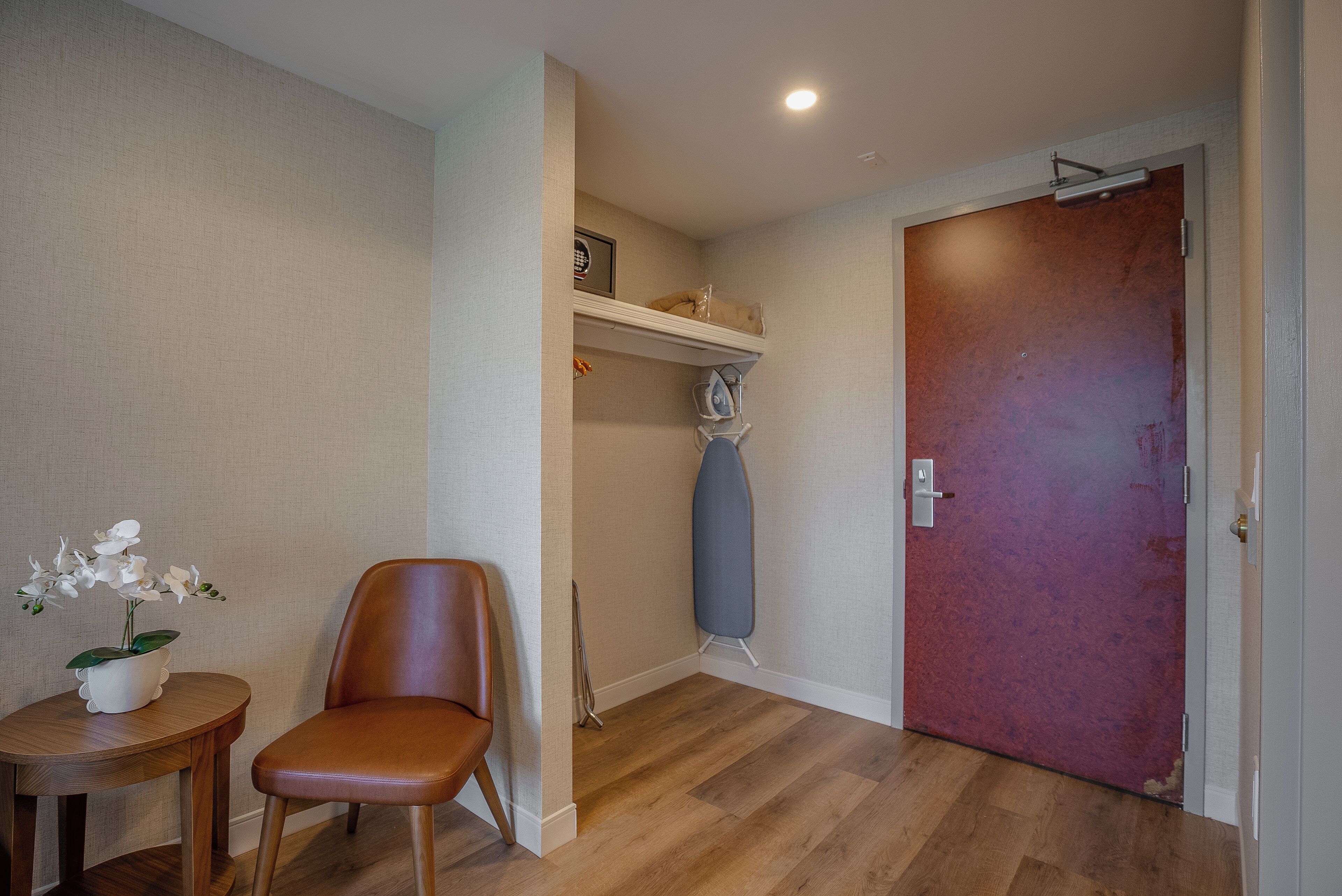 Ein Zimmer mit Holzboden, einem braunen Ledersessel, einem kleinen Tisch mit Blumentopf, einer roten Tür und einem Regal mit gerahmtem Bild und Bügelbrett in Blau.