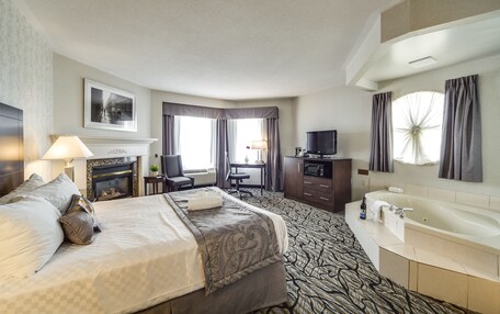 A hotel room with a large bed, a fireplace, a desk with a chair, a TV, a bathroom with a bathtub, and a large window with curtains.