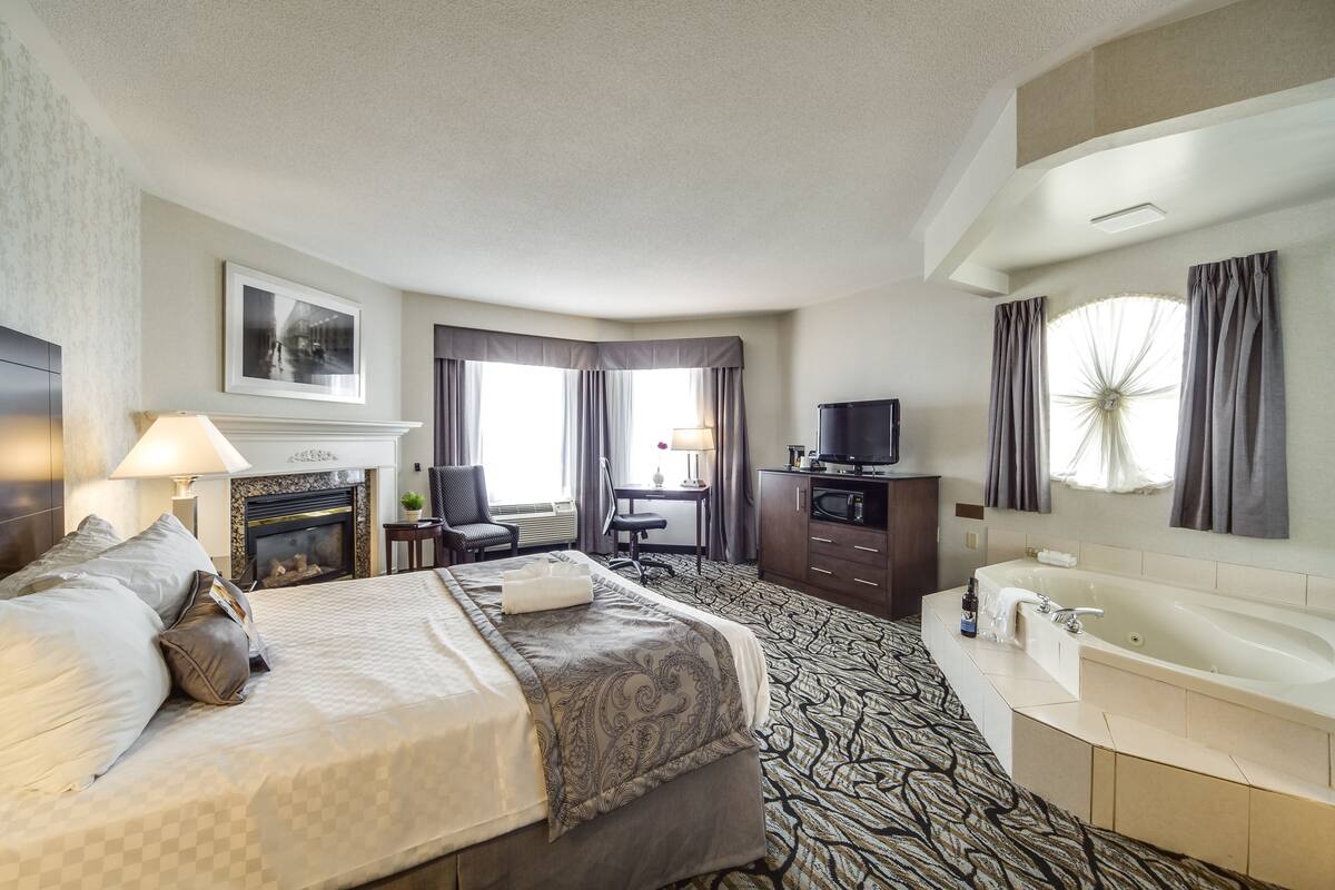 Une chambre d’hôtel dotée d’un grand lit, d’une cheminée, d’un bureau avec une chaise, d’une télévision, d’une salle de bain avec baignoire et d’une grande fenêtre avec des rideaux.