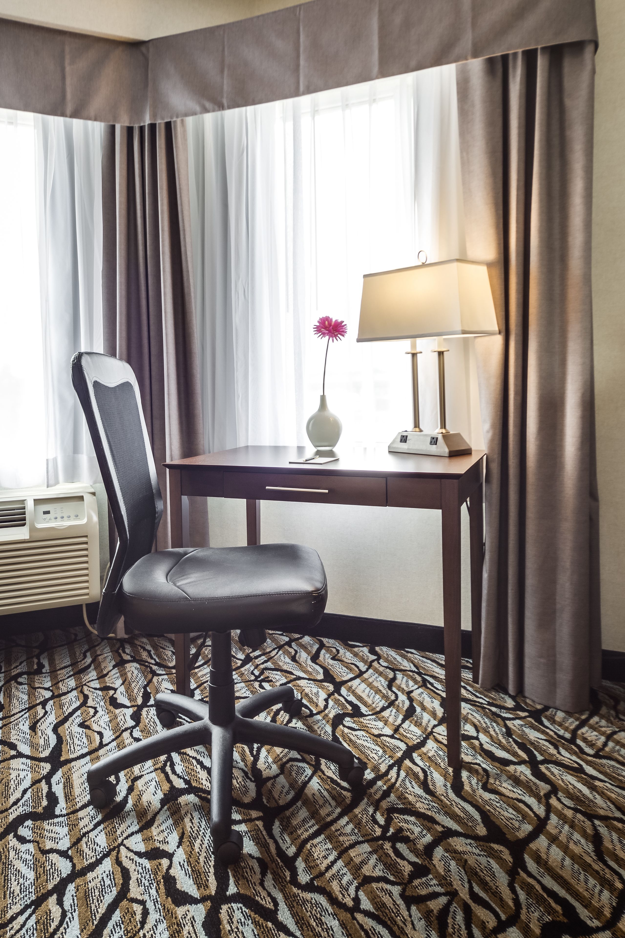 Ett hotellrum med ett skrivbord, en stol, en lampa, en luftkonditioneringsanläggning och en mönstrad matta.