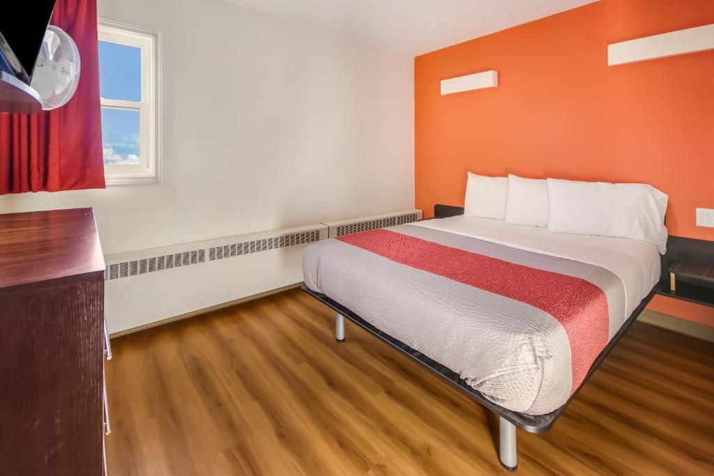 A hotel room with a bed, a wooden dresser, a window with curtains, and a wall heater.