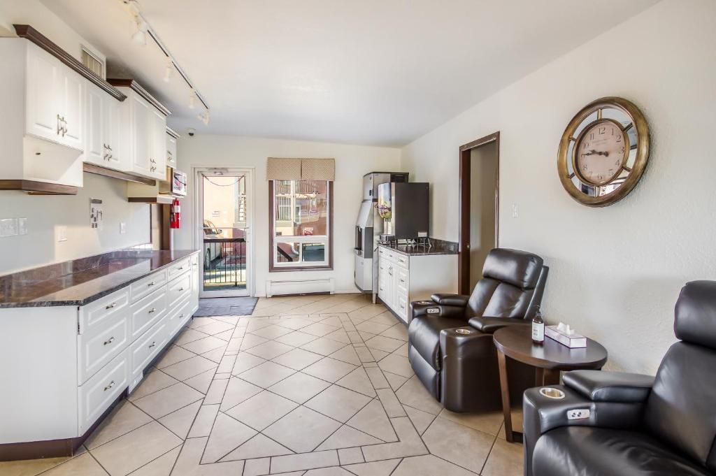 A modern kitchen with white cabinets and a marble countertop, a dining area with a black leather sofa and a small table, and a large wall clock.