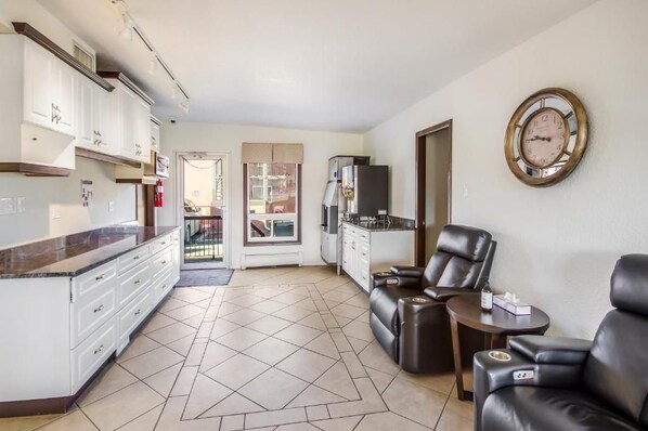 A modern kitchen with white cabinets and a marble countertop, a dining area with a black leather sofa and a small table, and a large wall clock.