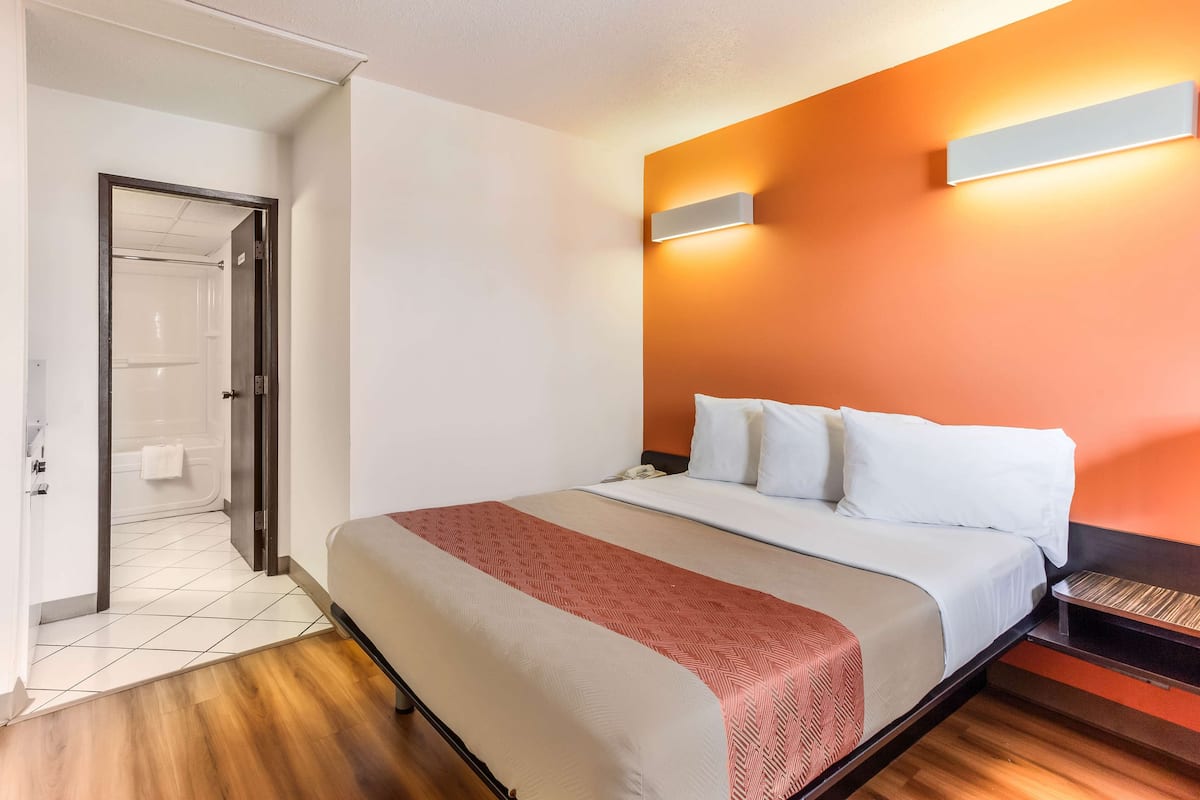 Una habitación de hotel con cama, mesita de noche, lámpara fijada a la pared y puerta que da a un baño.