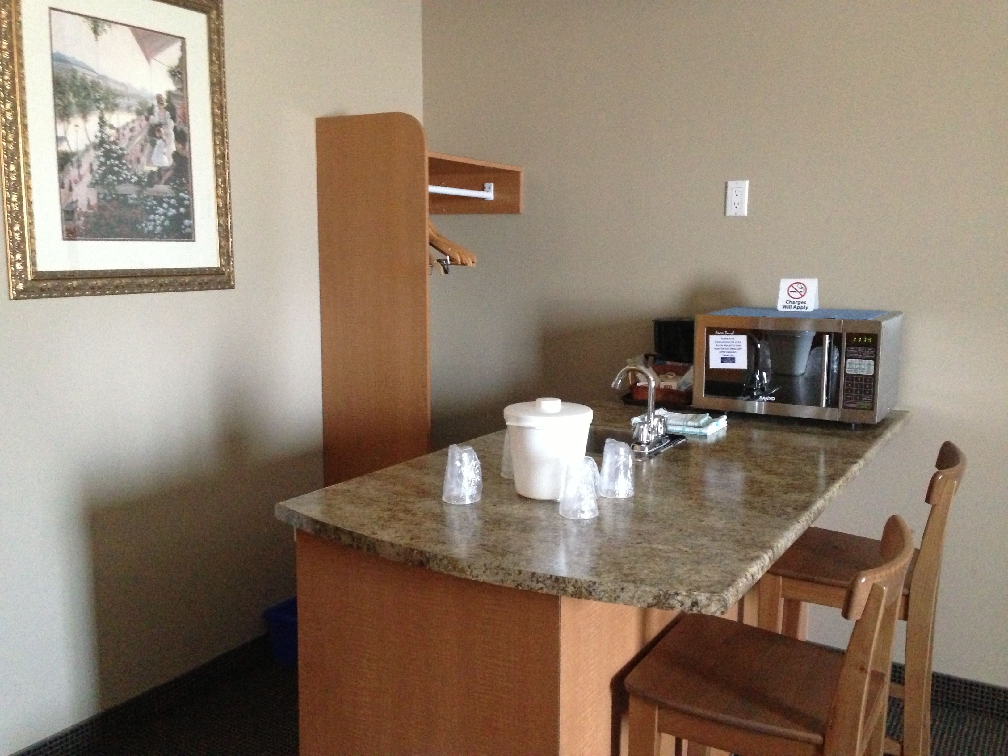 A small kitchenette with a microwave, a coffee maker, and a sink. There is a framed picture on the wall and a wooden chair.