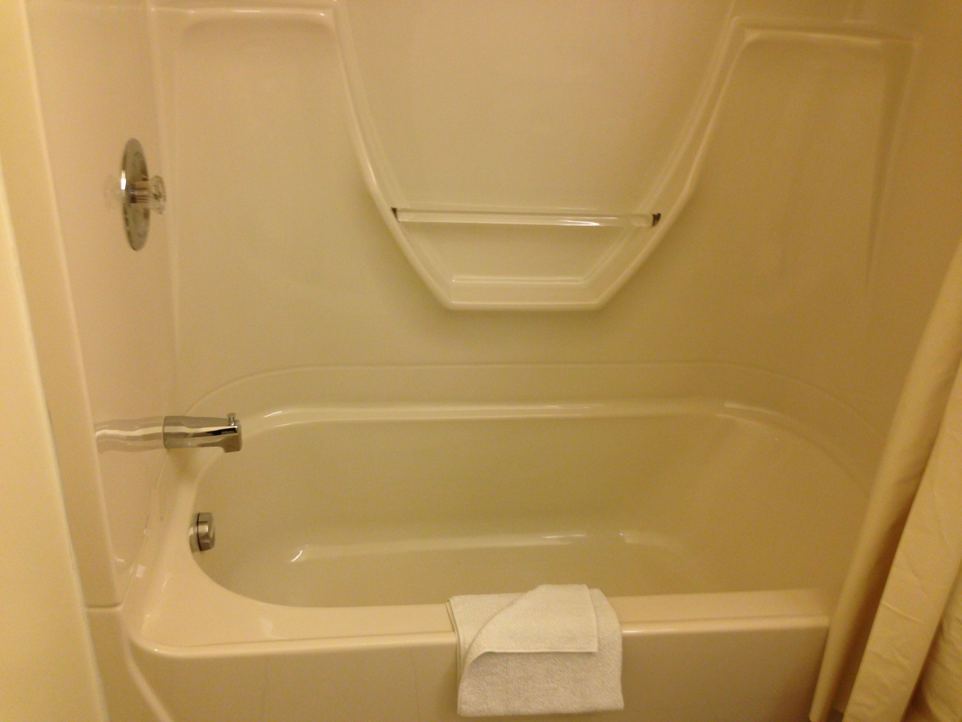 A standard bathtub with a towel on the side, a shower control panel, and a clear glass shelf above the tub.