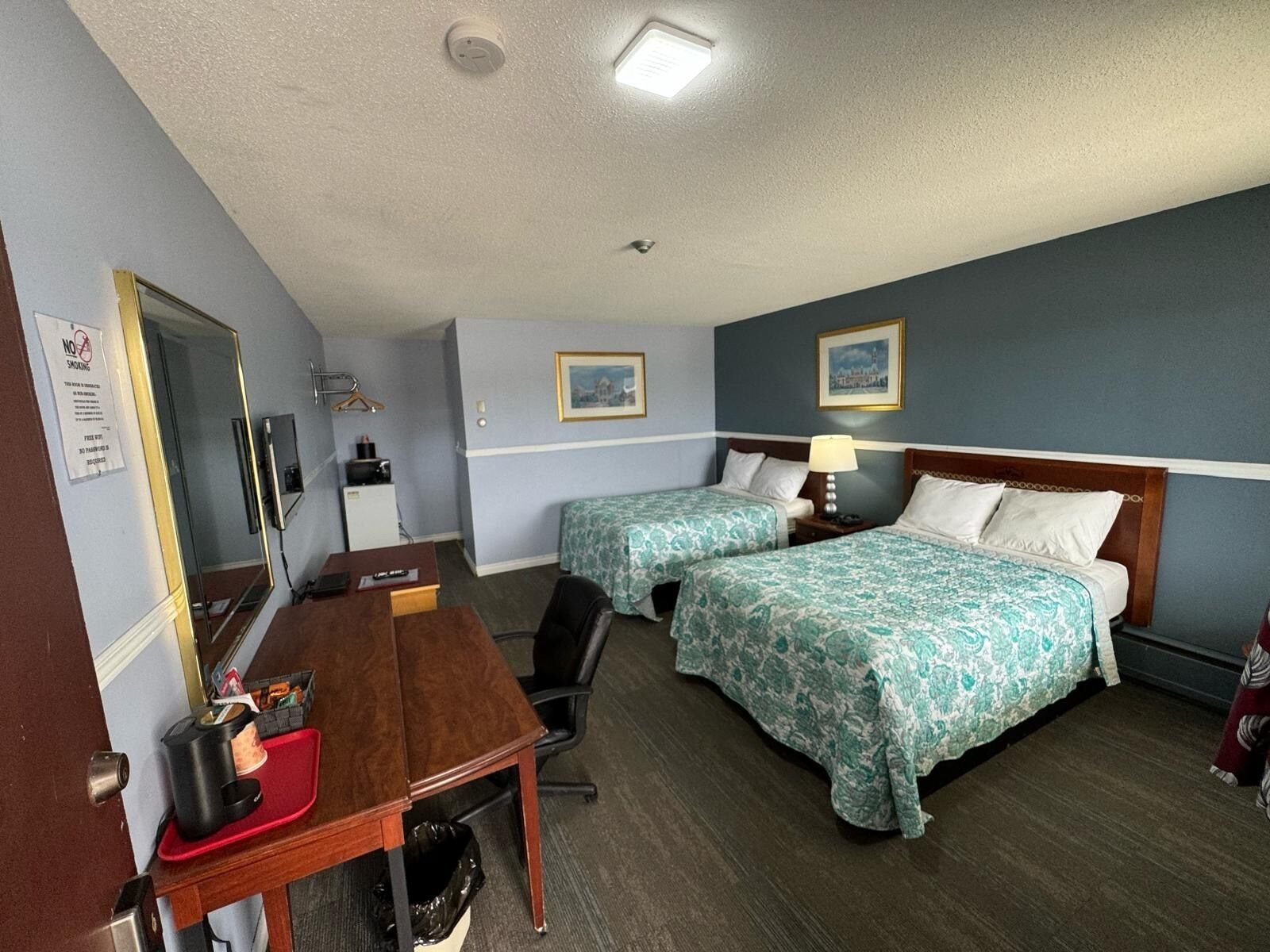 A hotel room with two beds, a desk, a chair, a mirror, a TV, and a trash bin.