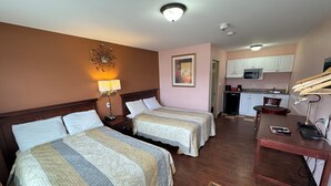 Habitación de hotel con dos camas, un escritorio, una silla, una kitchenette pequeña y un baño visible a través de una puerta abierta.